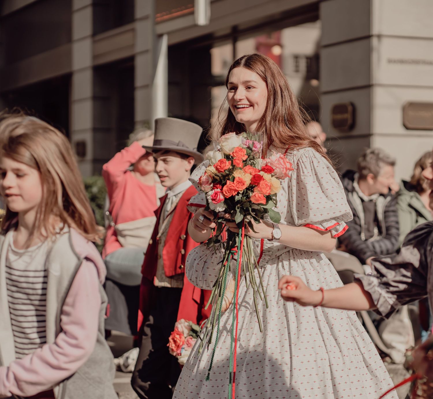 Young woman in a vintage dress smiles while holding a bouquet of pink roses at a street parade.