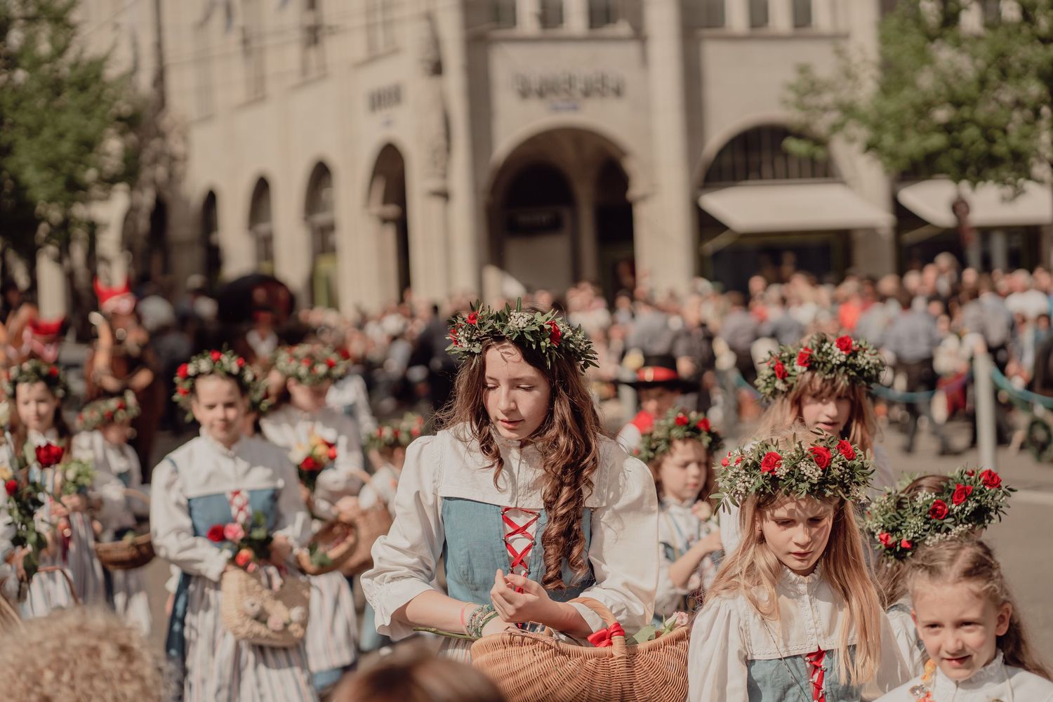 Women and girls in white folk costumes with red floral wreaths march in traditional cultural parade.