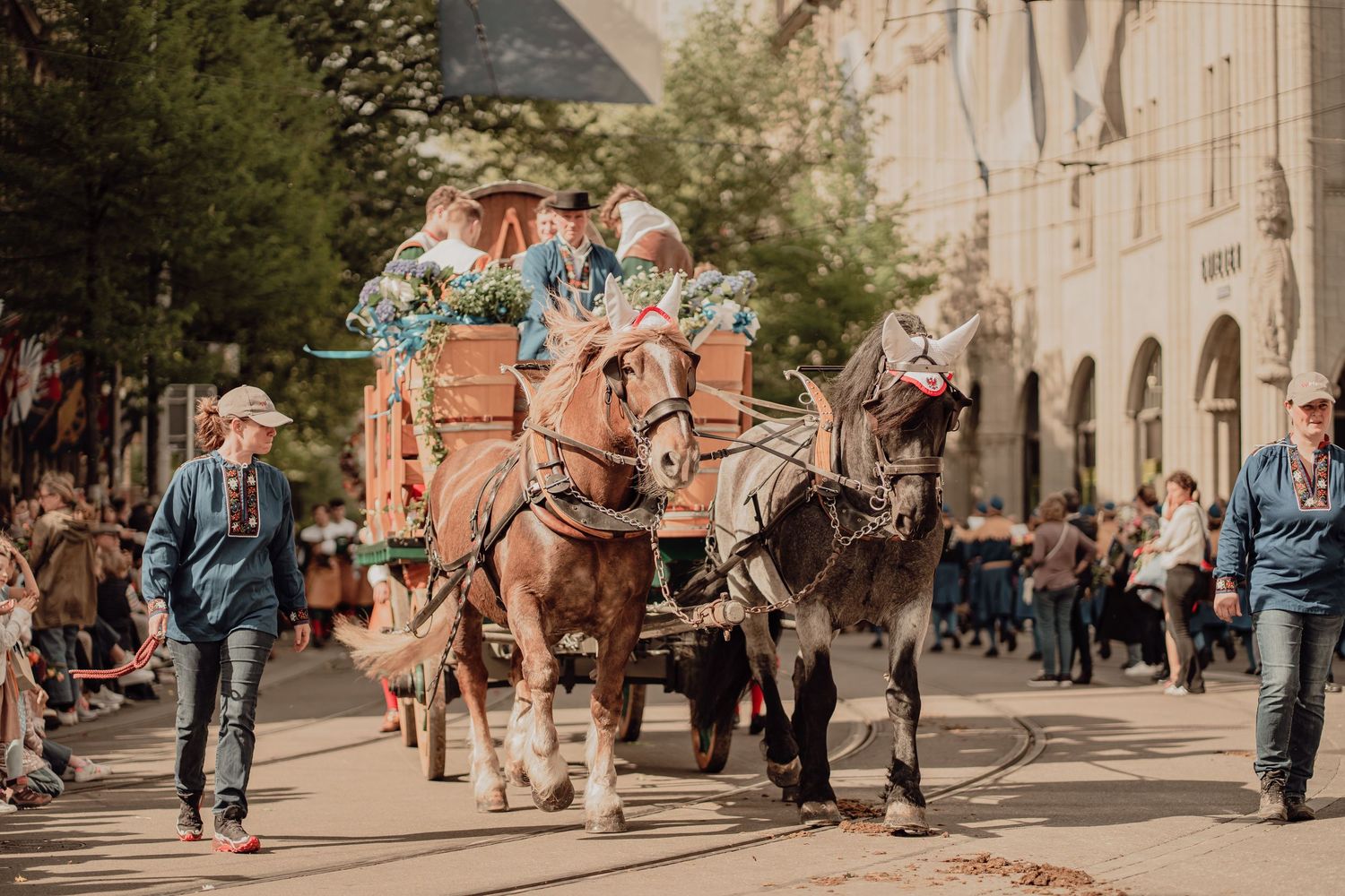 A horse-drawn cart decorated with flowers moves through a crowded traditional European street parade.