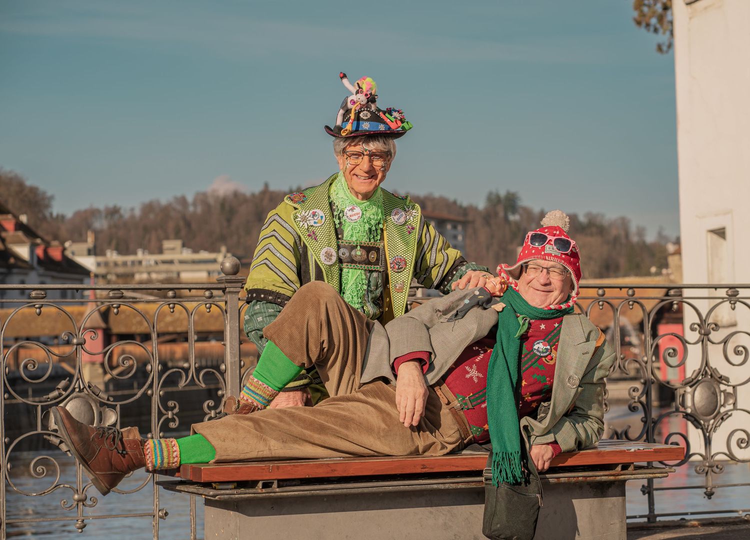 Two carnival performers in colorful costumes playfully pose on a railing with a scenic European town backdrop in sunny weather.