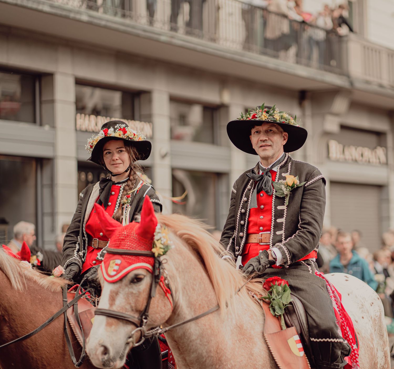 Two riders in traditional red and black costumes with decorated hats ride horses in a festive parade.
