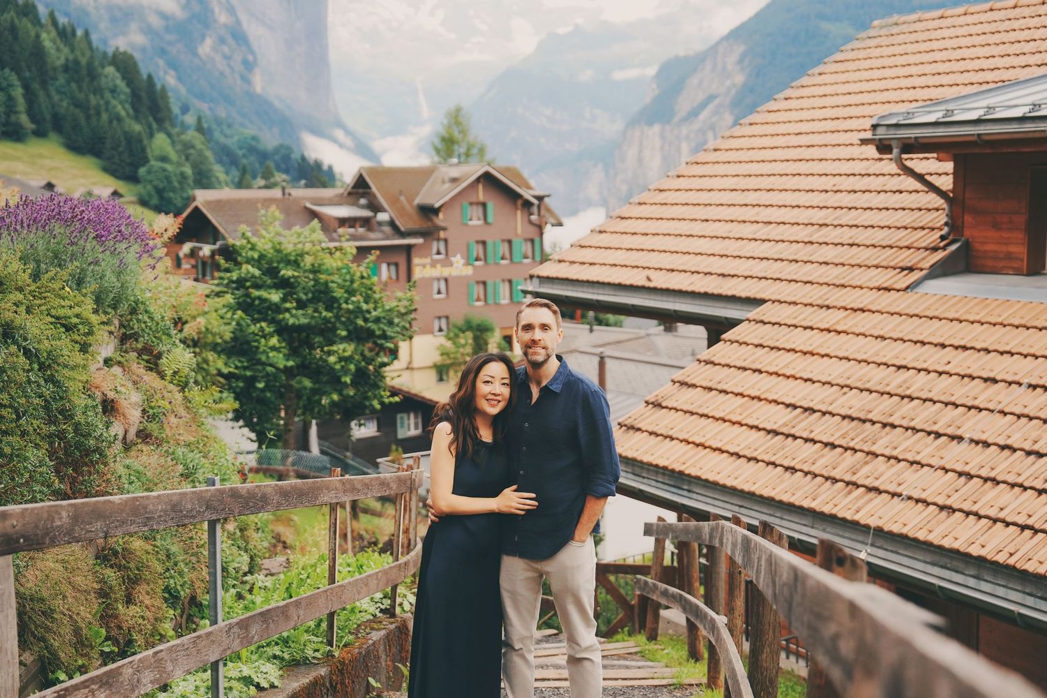 A smiling couple embraces on a wooden balcony overlooking a picturesque Swiss mountain village.
