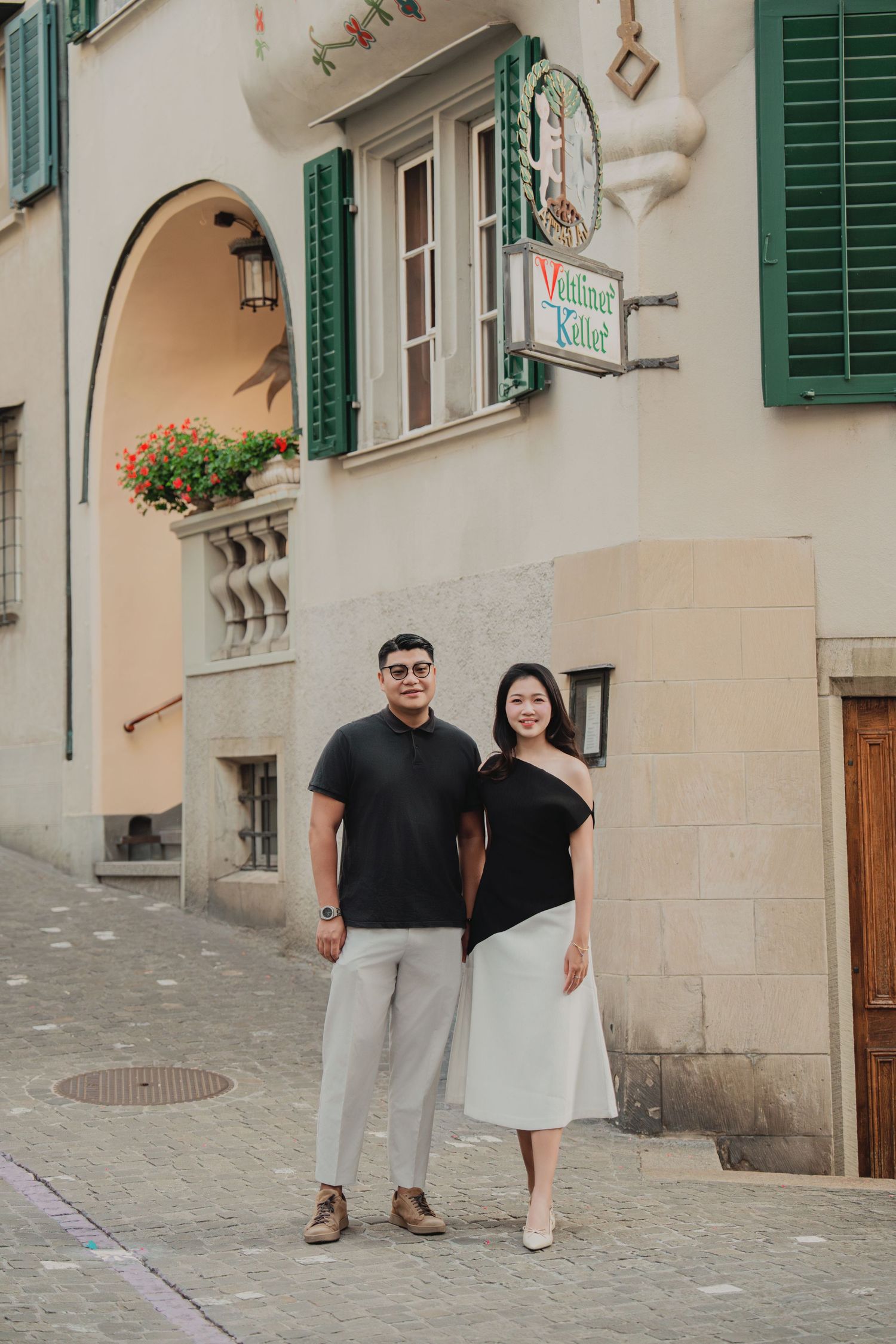 Couple posing together on a cobblestone street beside a historic European building with green shutters.