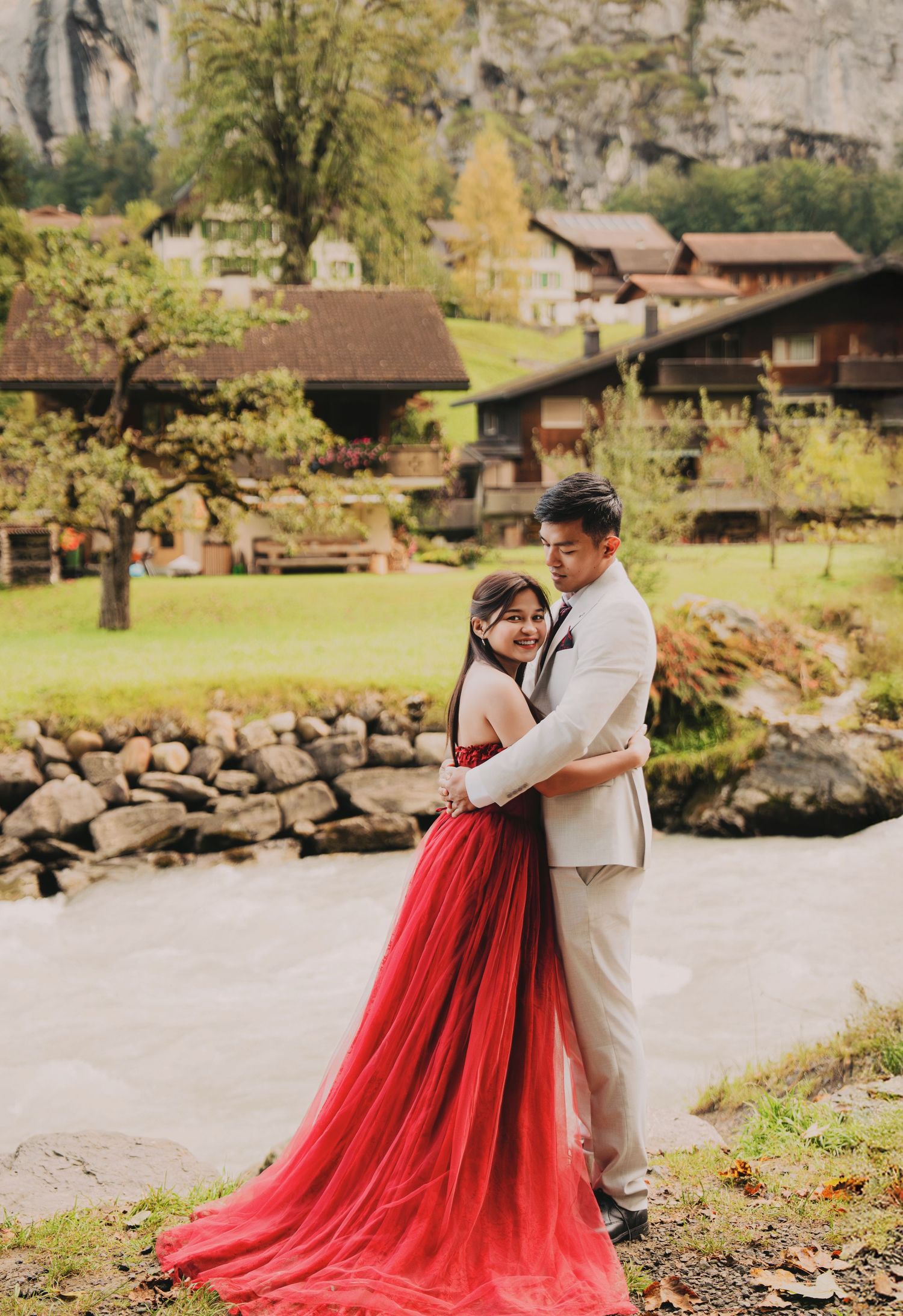 Couple in red wedding gown and beige suit pose romantically by river in Swiss mountain village.