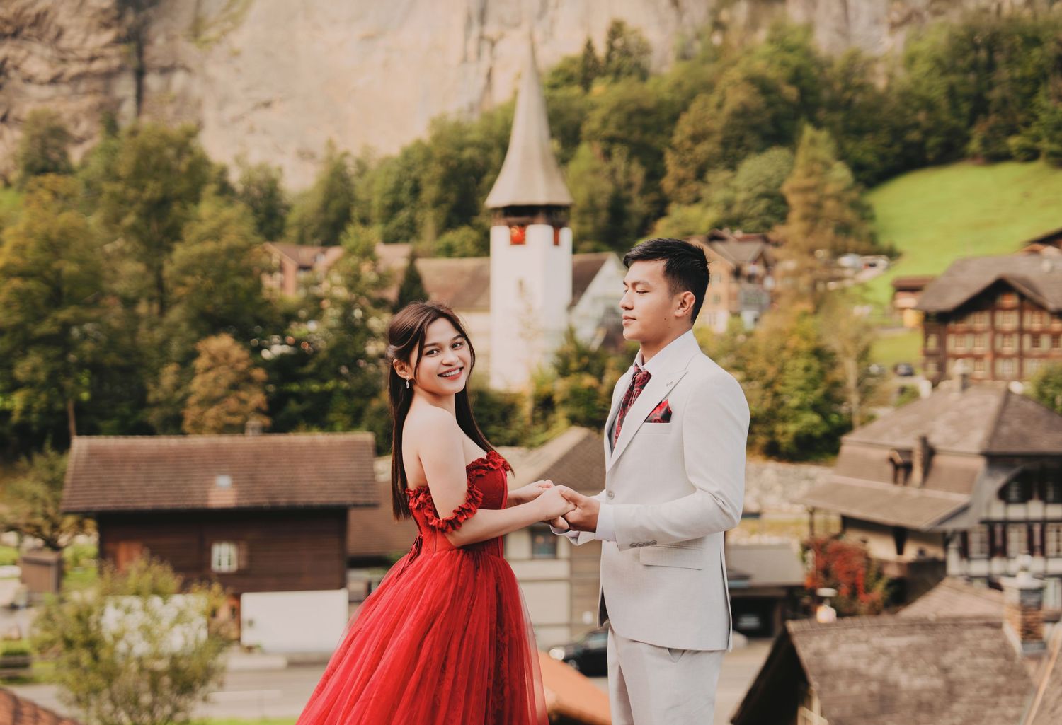 Smiling couple holding hands with a picturesque Swiss village church and rocky cliffs behind them.