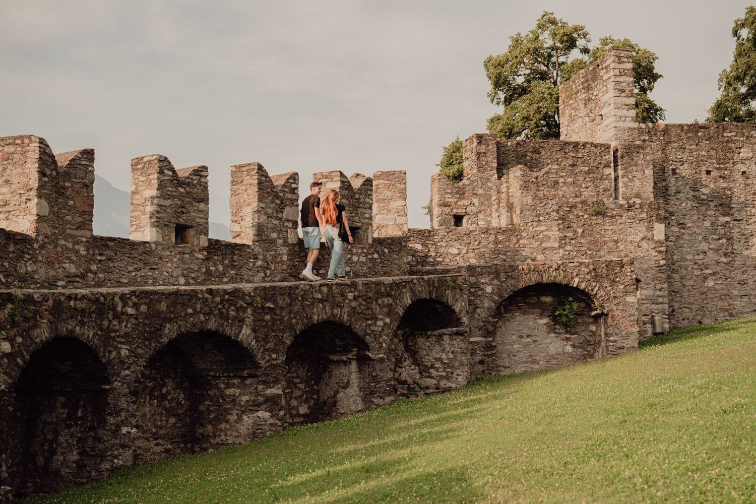 A romantic couple sitting atop a medieval castle battlement wall overlooking a lush green courtyard.