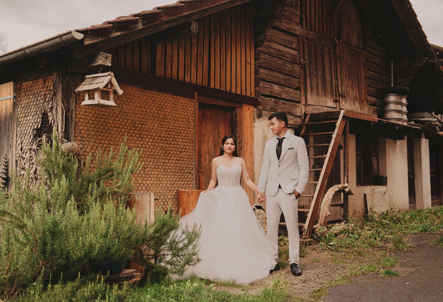 Elegant couple in wedding attire holding hands before a rustic wooden Swiss barn surrounded by lush greenery.