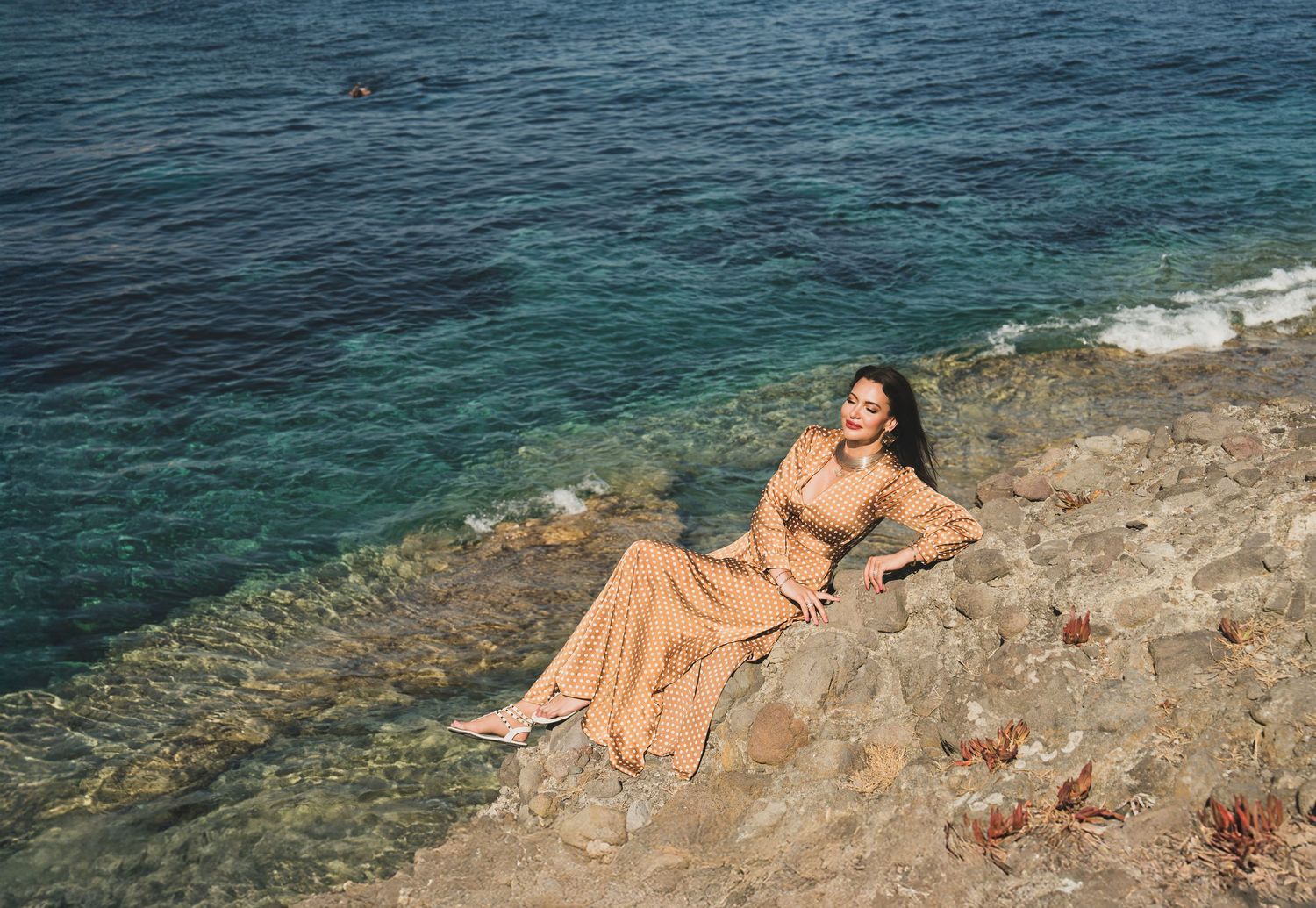 Woman in beige maxi dress reclining on sun-drenched rocks beside clear turquoise sea.