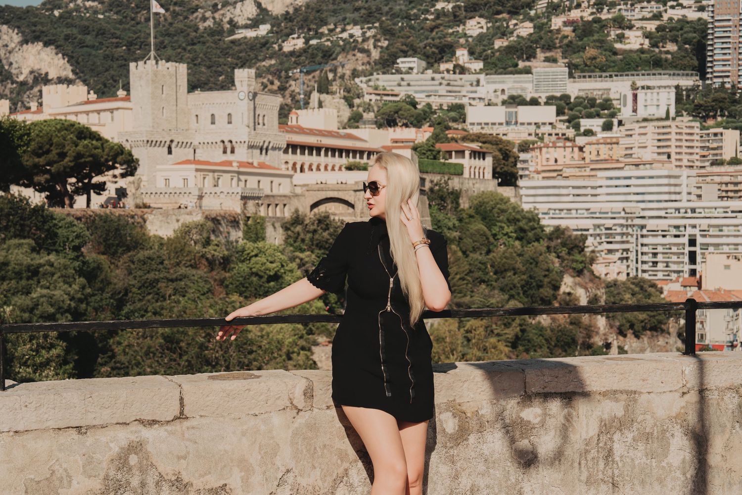 Blonde woman in black dress overlooking Monaco Palace from scenic stone terrace.