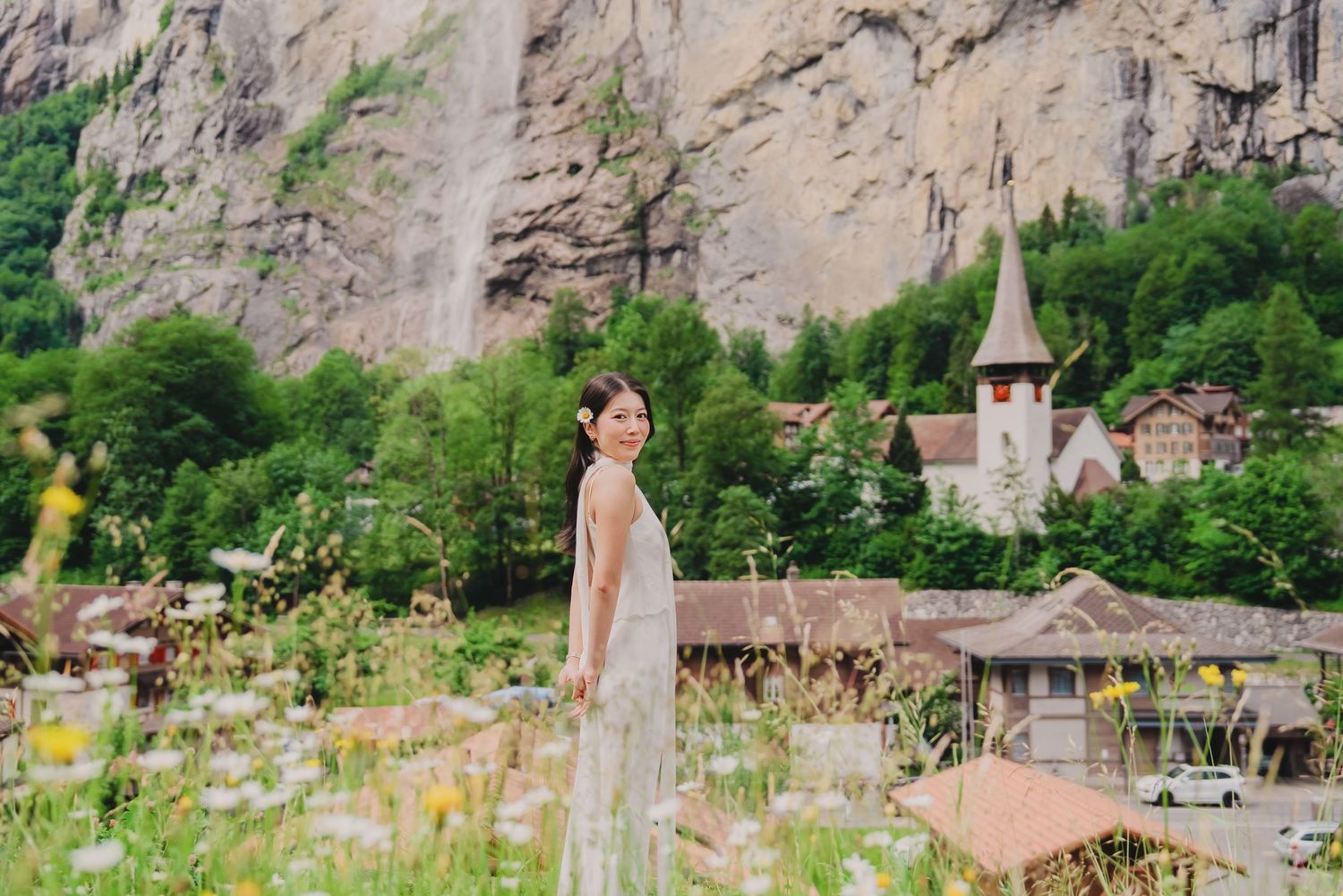 Woman in floral dress standing in wildflower meadow with Lauterbrunnen village and waterfall in background.