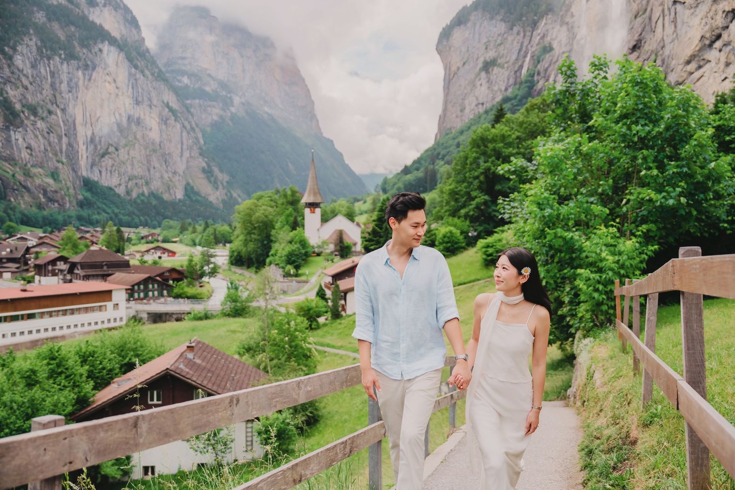 Couple holding hands walking on a bridge with Lauterbrunnen valley village and Swiss Alps in background.
