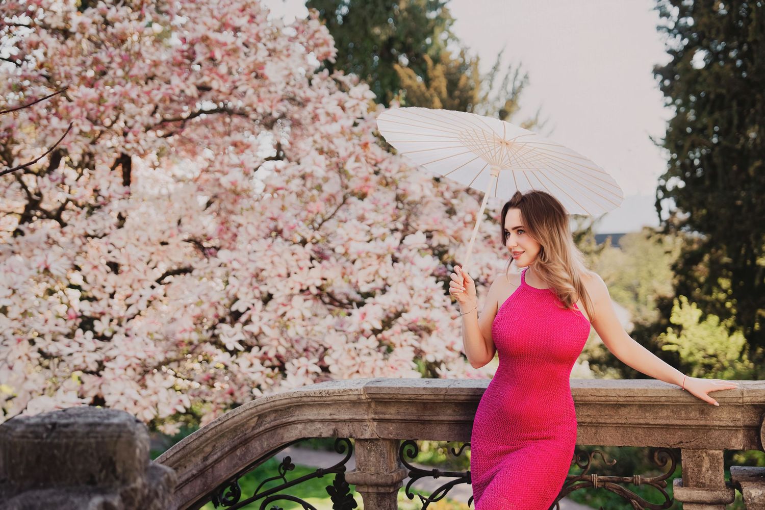 Woman in pink dress holding white parasol poses by stone railing with blooming cherry blossom trees in background.