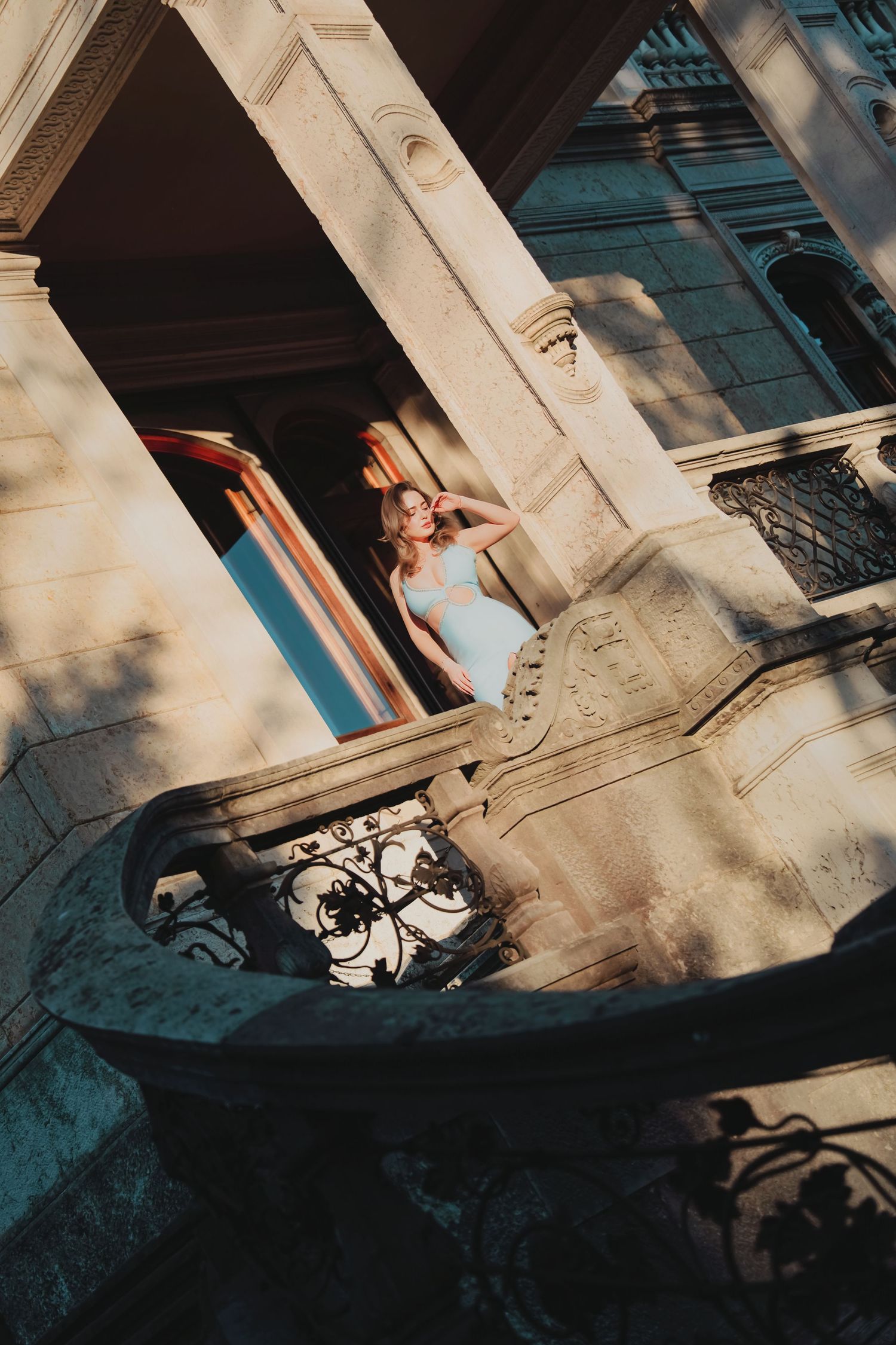 Woman in white dress poses on ornate stone balcony of historic building with columns in golden sunlight.