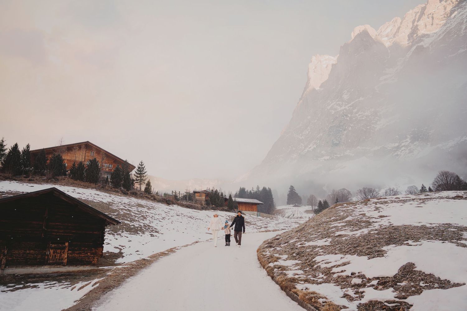 Family walking along snowy path past wooden chalet with foggy mountain landscape in winter.