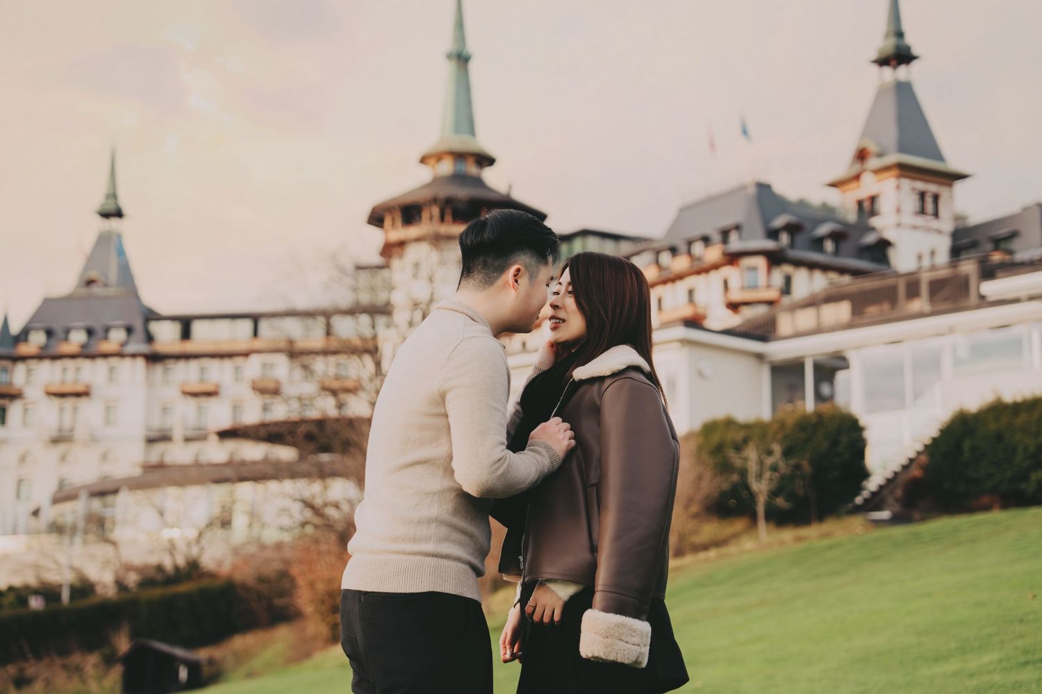 Young Asian couple sharing a kiss on green lawn outside European castle hotel.