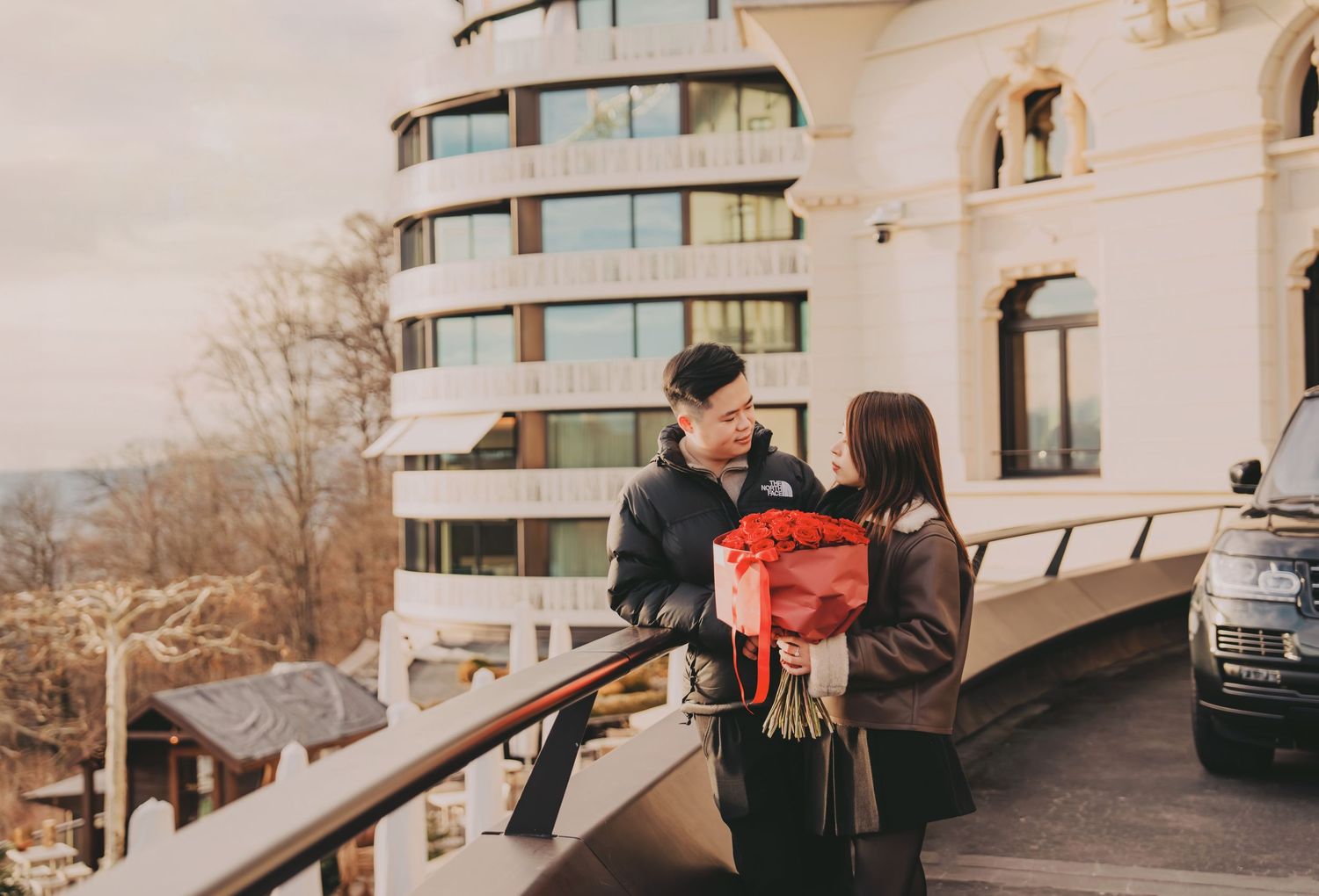 Asian man presenting a bouquet of red roses to woman outside a luxury hotel building.