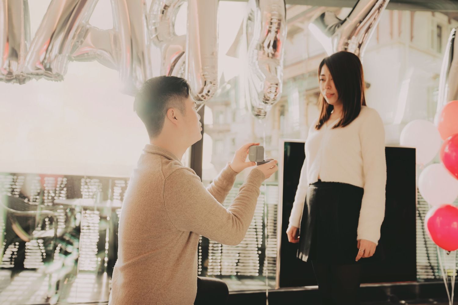 Man kneeling to propose to woman in romantic balloon-decorated indoor setting.