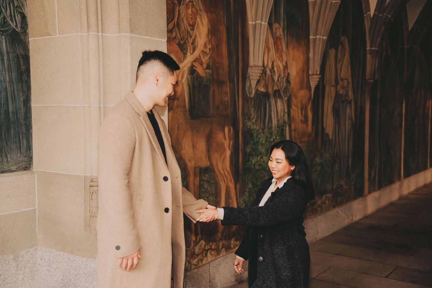 Couple holding hands in a corridor adorned with classical painted murals on stone walls.