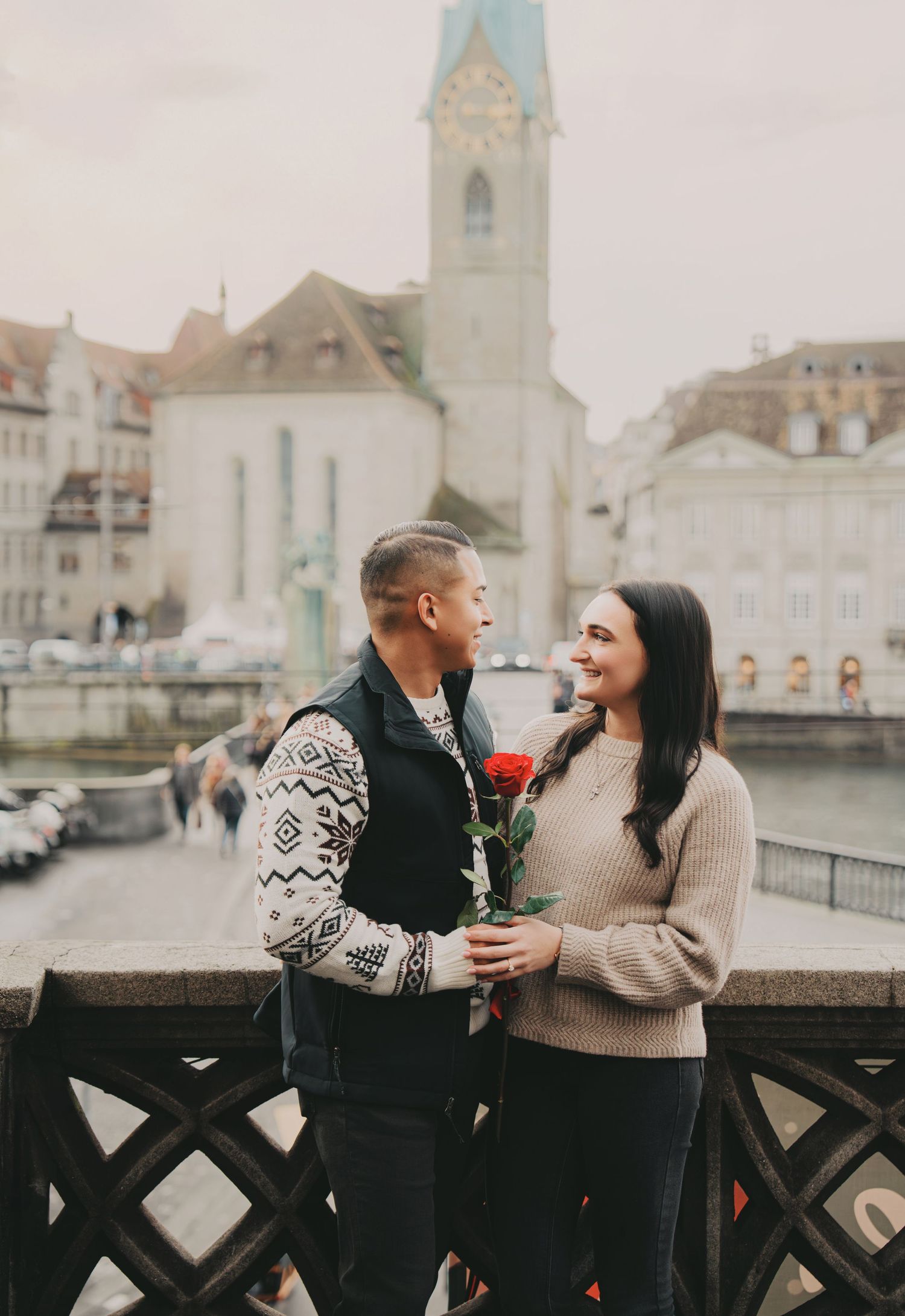 Romantic couple sharing a red rose on a bridge with Fraumunster Church in Zurich background.
