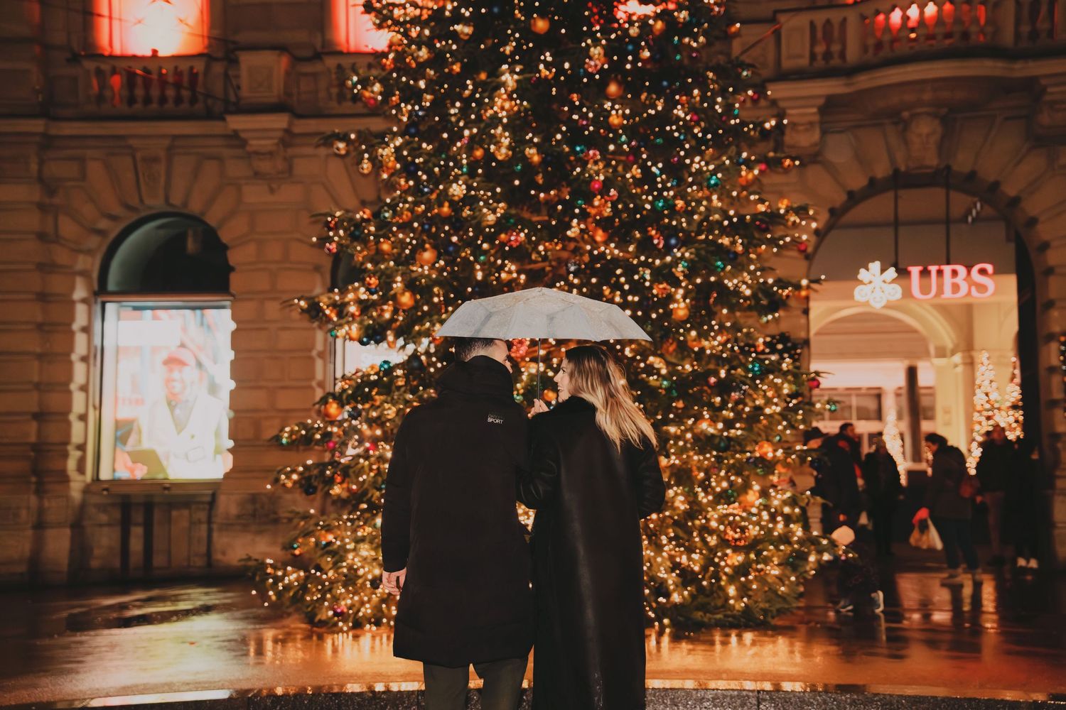 Couple sharing a kiss under an umbrella beside a grand illuminated Christmas tree in a city square.