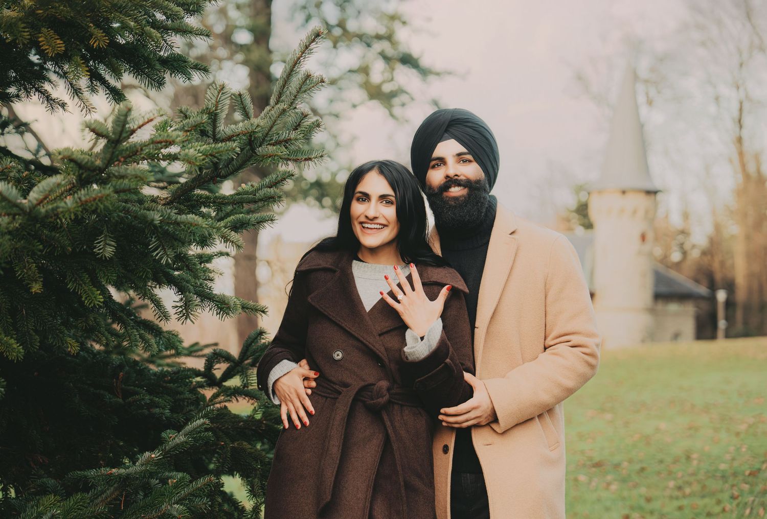 Smiling engaged couple posing by evergreen tree, showing off ring.