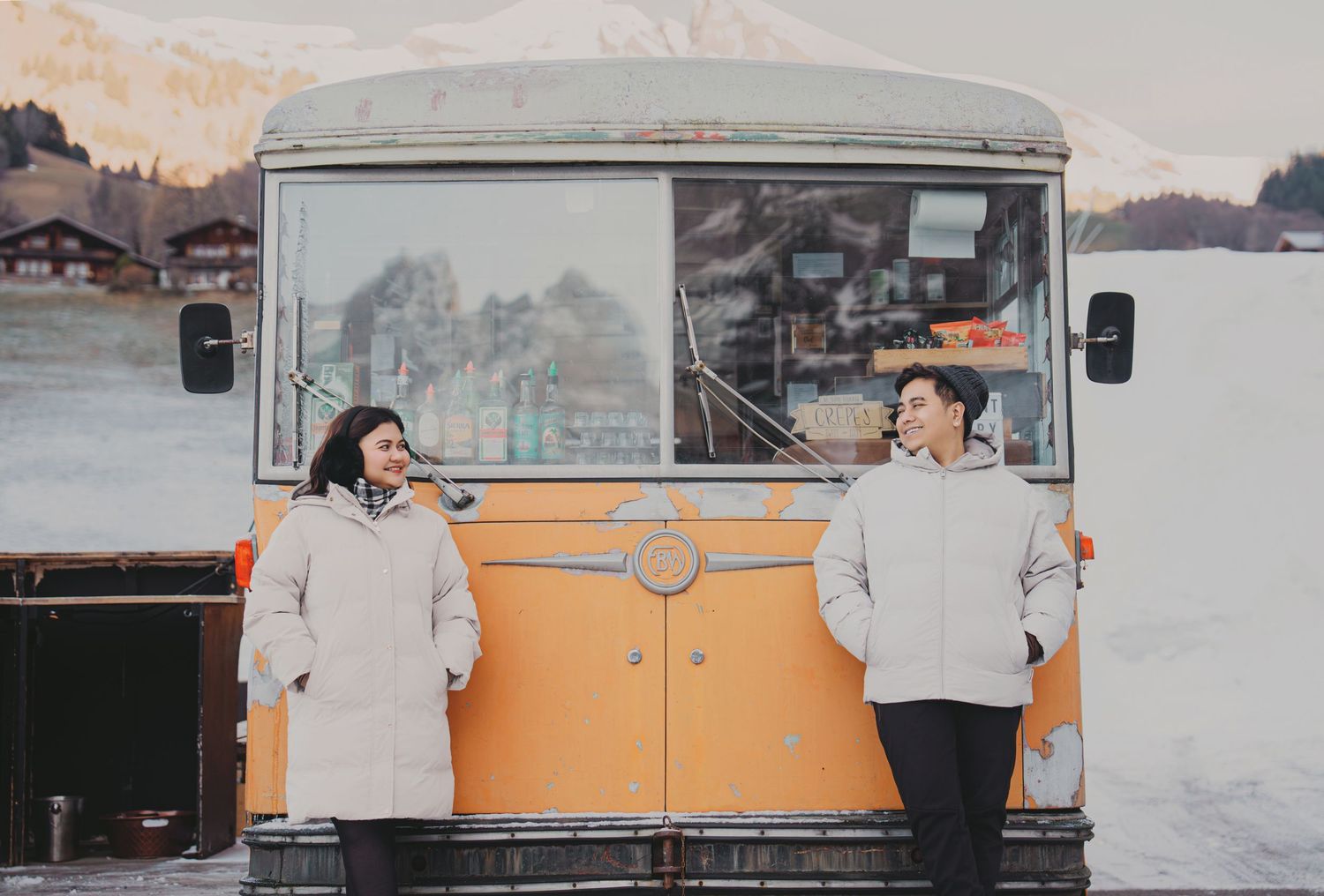 Couple in white winter coats standing apart beside a vintage orange food truck in snowy Alps.