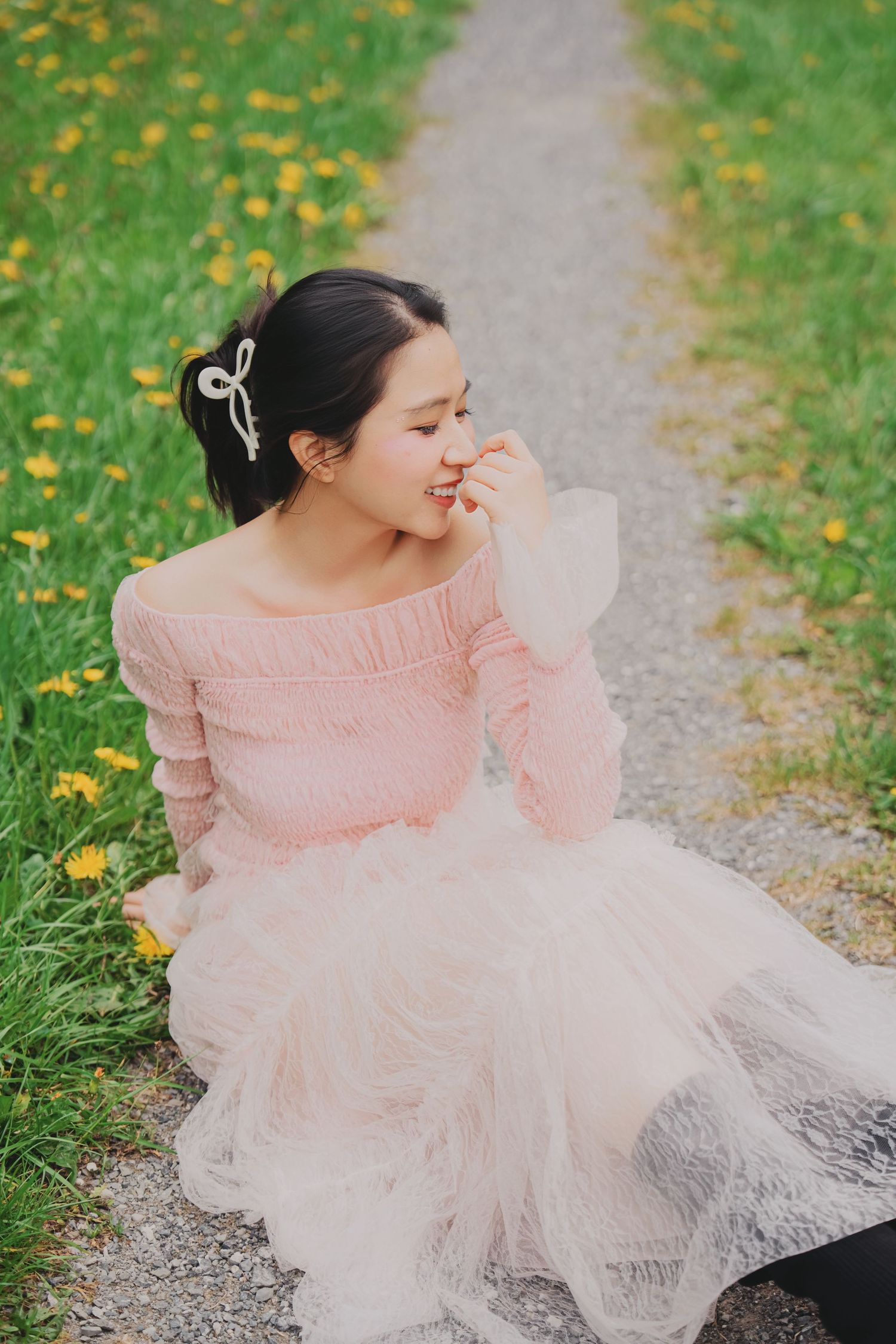 Young woman in pink off-shoulder dress sitting on garden path among dandelions with bow hair accessory.