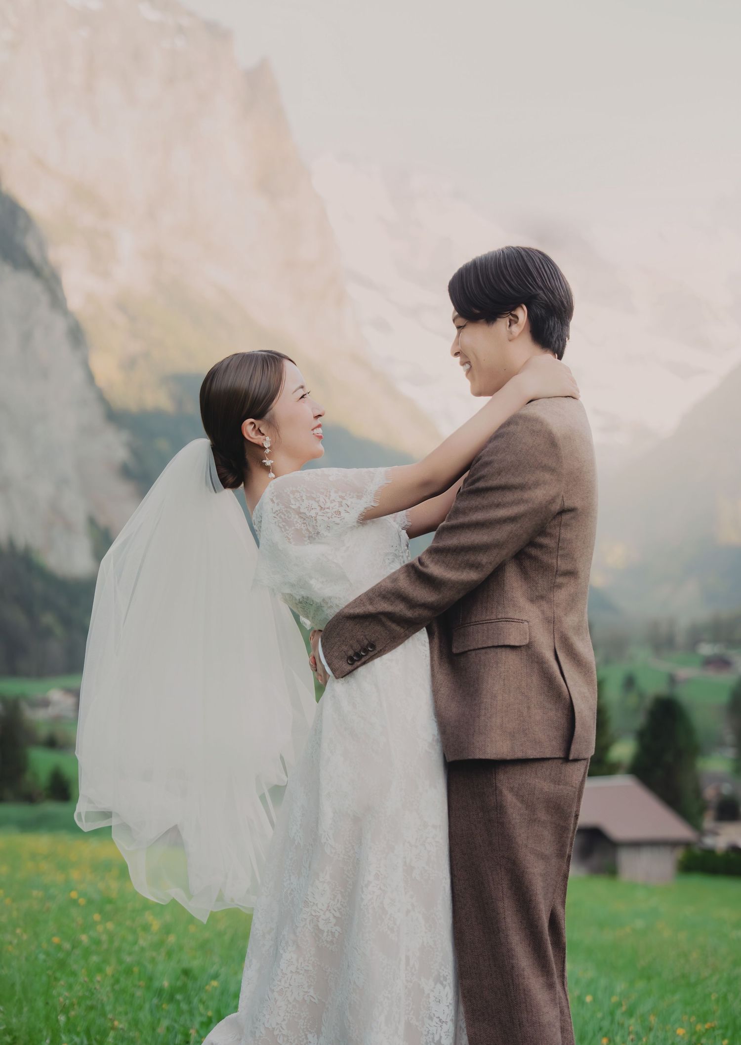 Bride and groom laughing together in a green Swiss alpine meadow with dramatic mountain cliffs in the background.