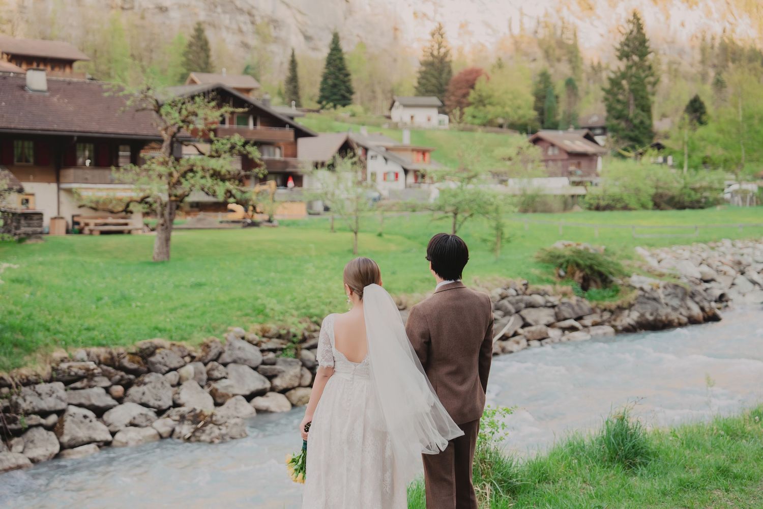 Bride and groom walking hand in hand along a peaceful riverside path in a lush Swiss Alpine village.