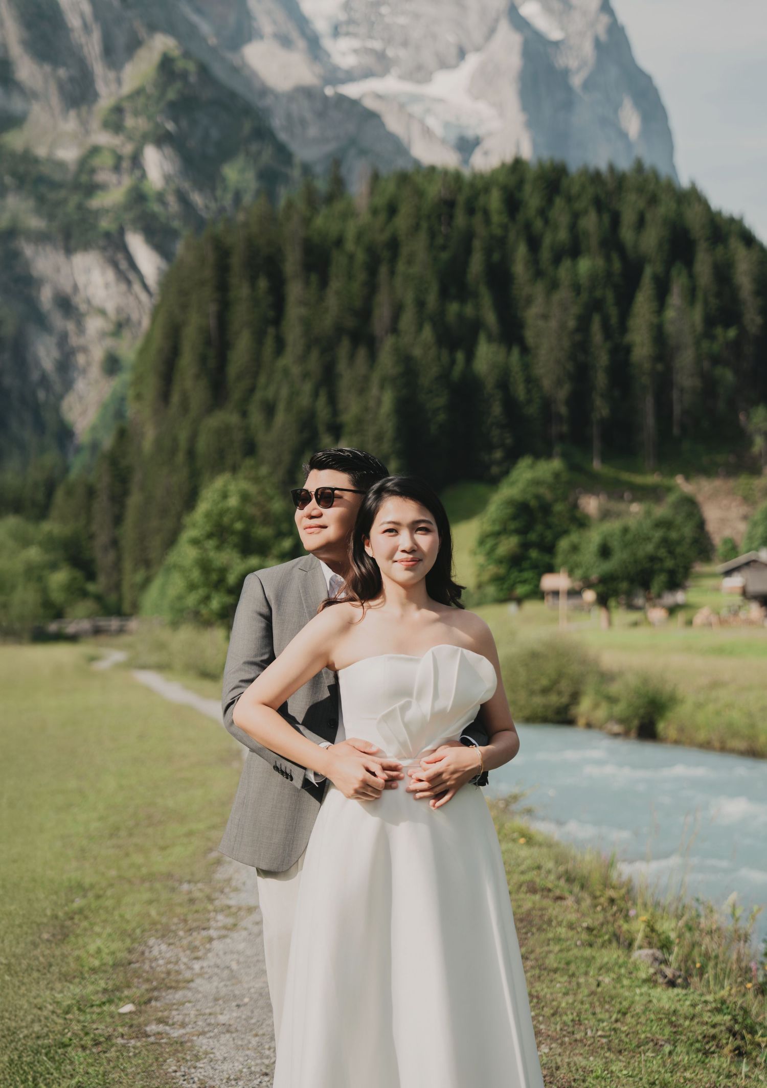 Groom standing behind bride in strapless white gown with majestic mountain and river backdrop.