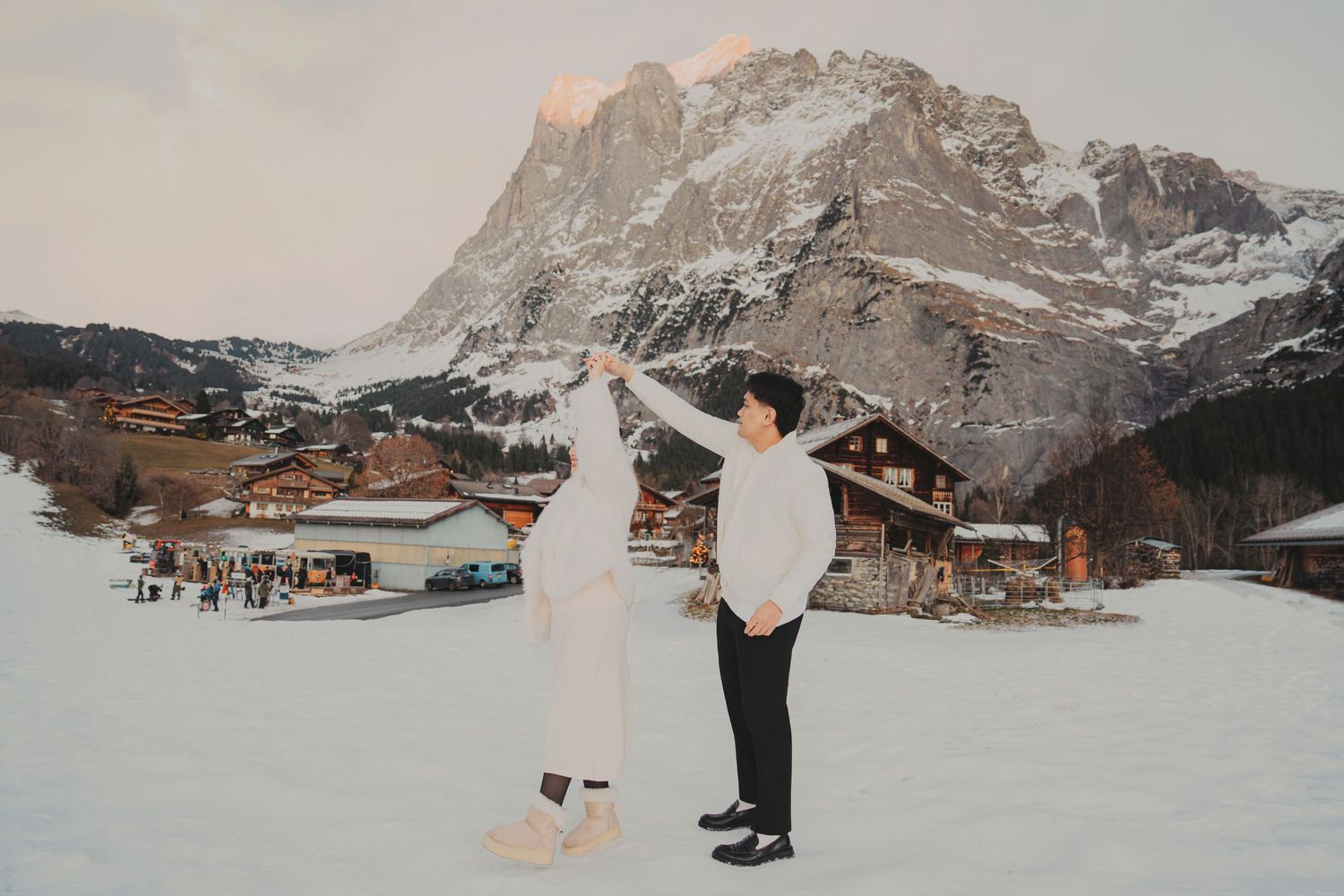 Couple dancing in snow with dramatic rocky mountain and Swiss alpine village in background at sunset.