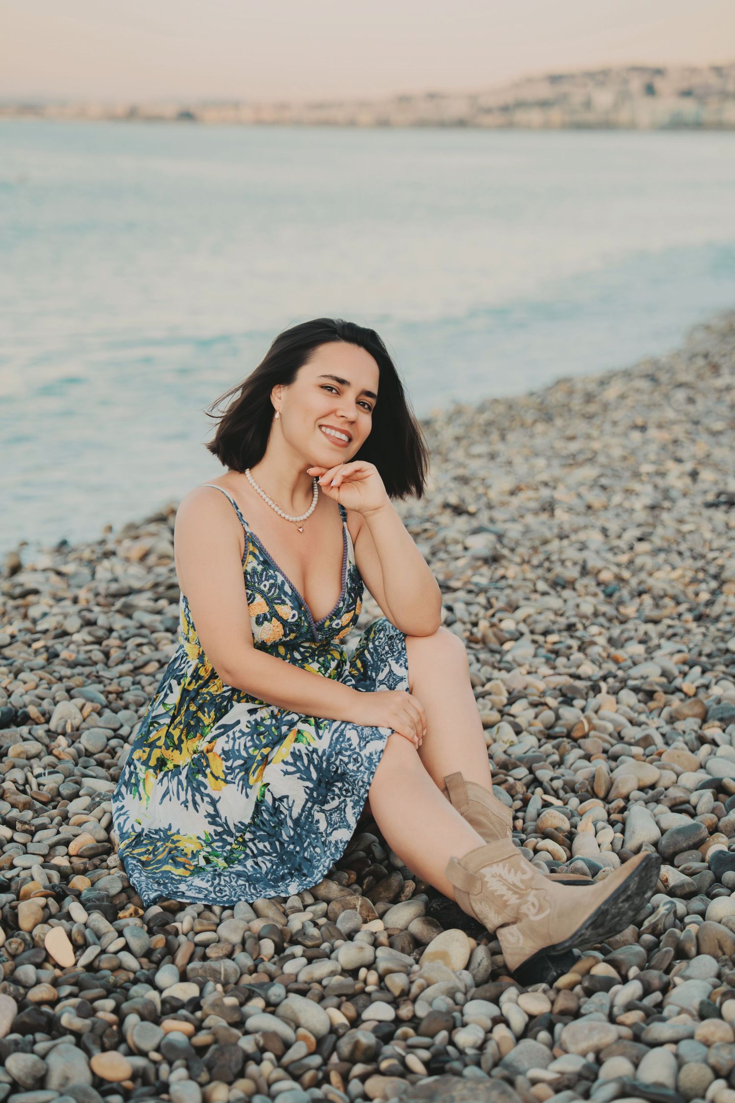 A cheerful woman in a floral dress and boots sits relaxed on a pebble beach.