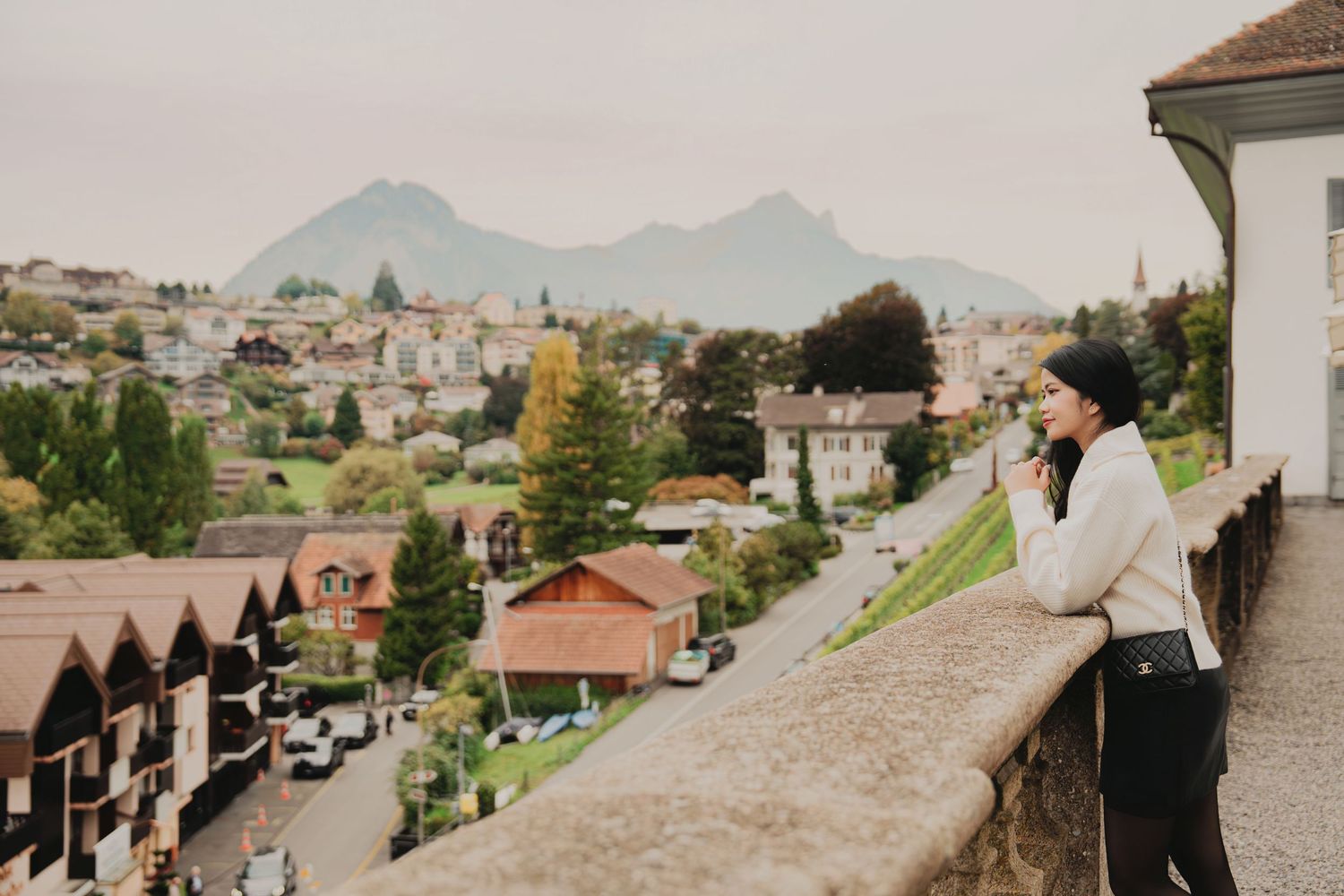 Woman in black beanie and white sweater admires view of Alpine town from stone balcony overlook.