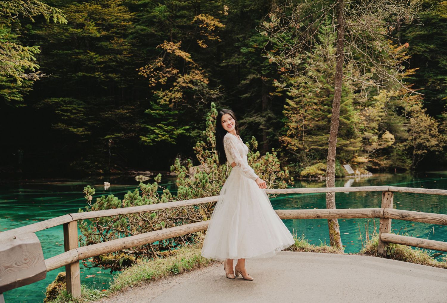 Young woman in white tulle dress posing on wooden deck overlooking a vibrant turquoise lake in forest.