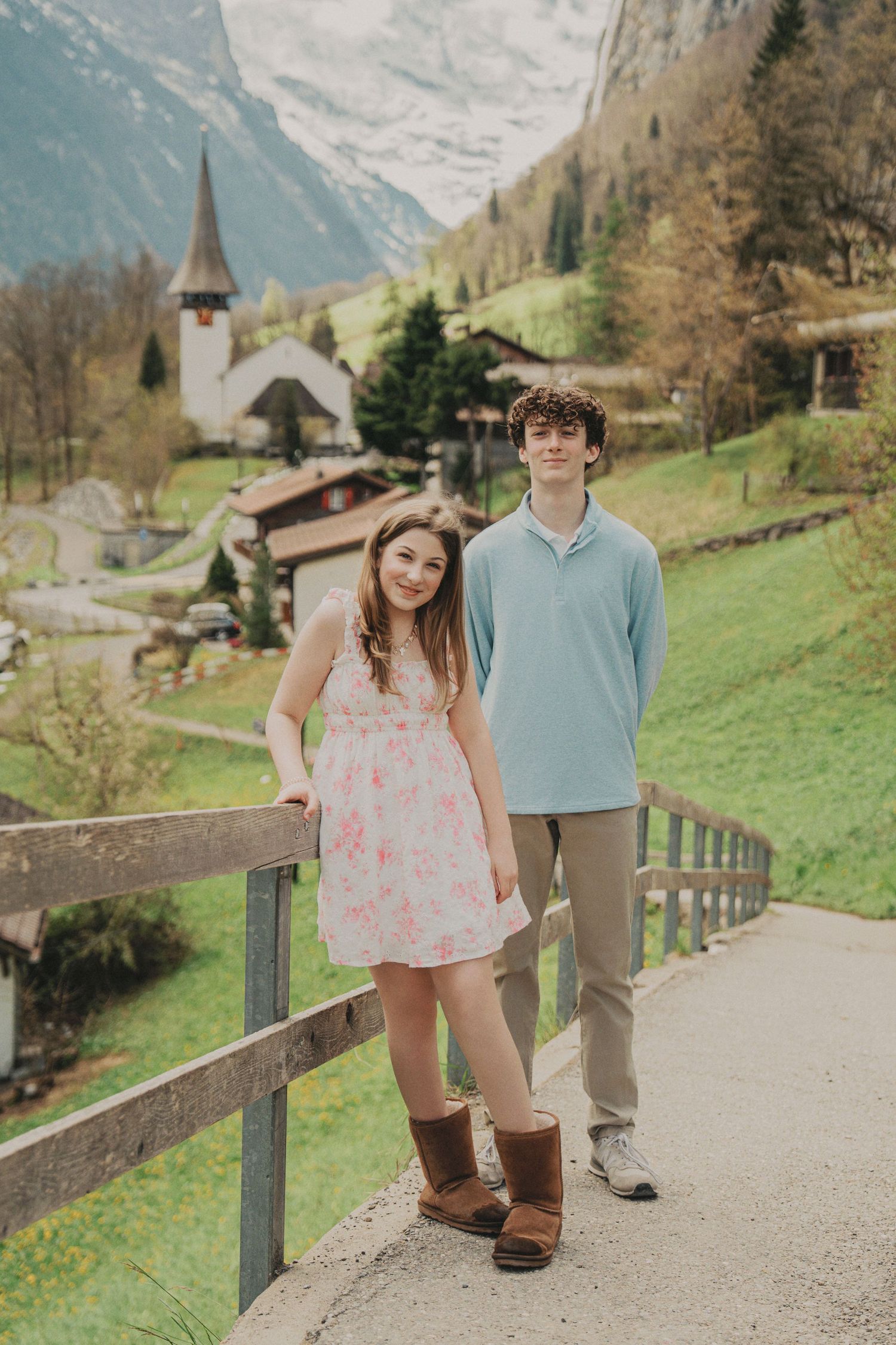 Teen boy and girl posing by a wooden fence with a Swiss alpine village church and mountains in the background.