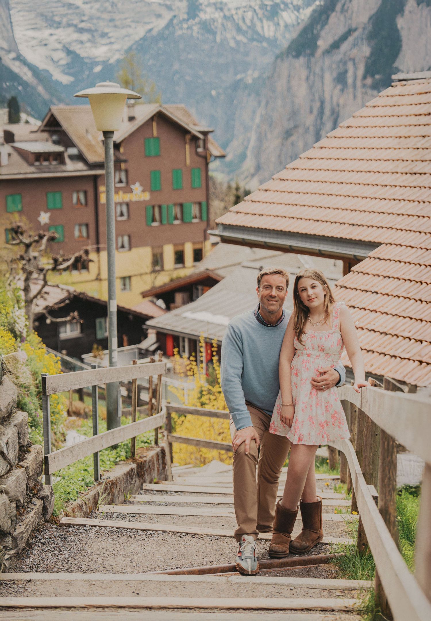 Father and daughter in floral dress posing together on wooden staircase in Swiss mountain village.