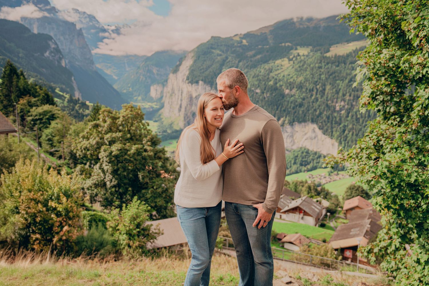 Couple embracing on a grassy hillside overlooking a picturesque Swiss mountain valley.