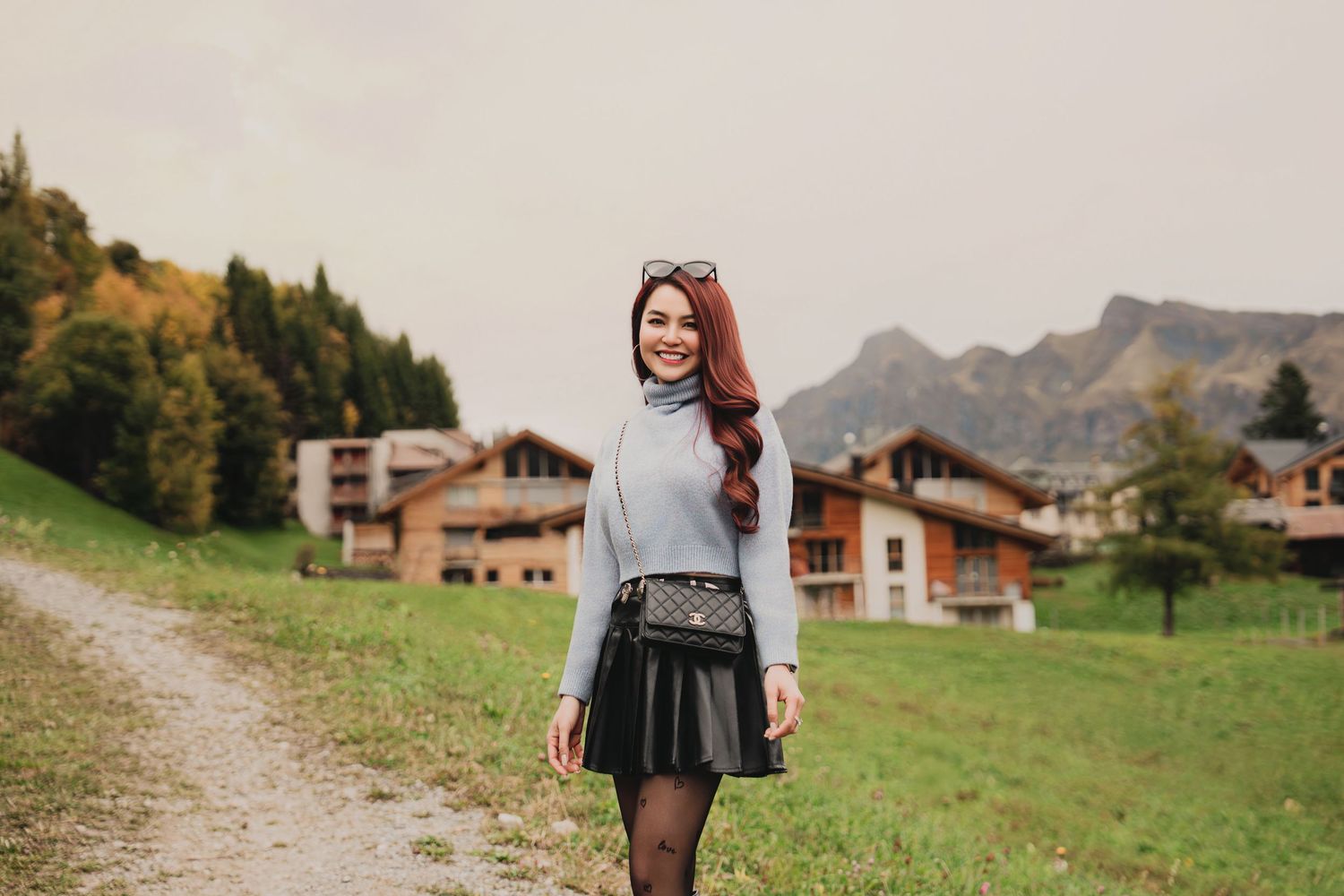 Smiling woman in grey sweater and black skirt walking on a path in a scenic alpine meadow.