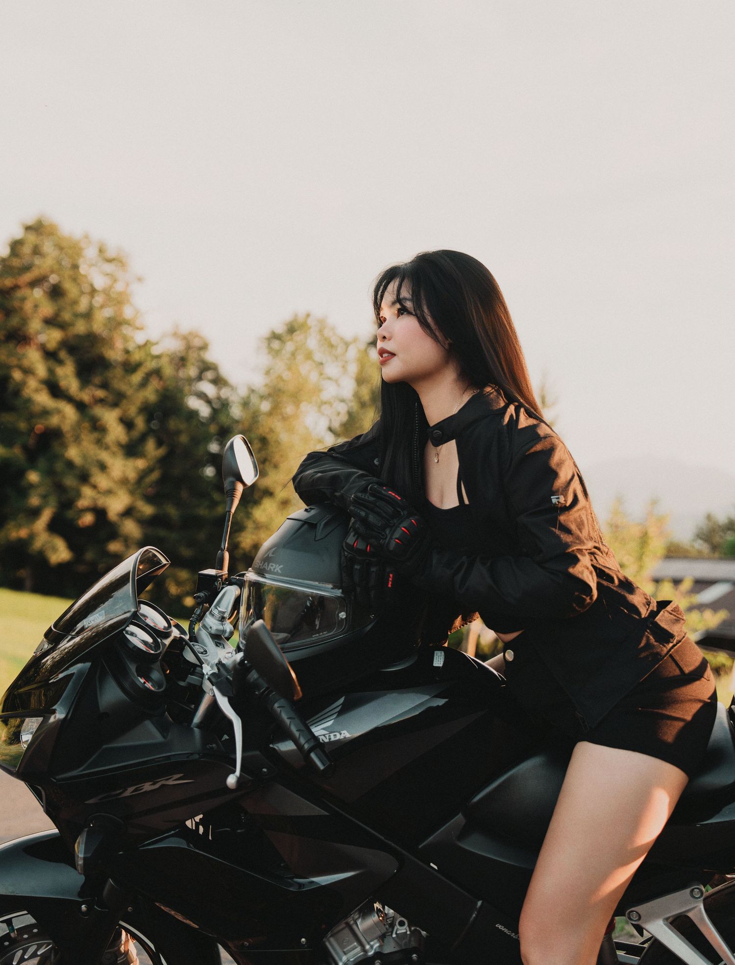 Woman in black jacket sitting on a black sport motorcycle outdoors at sunset.