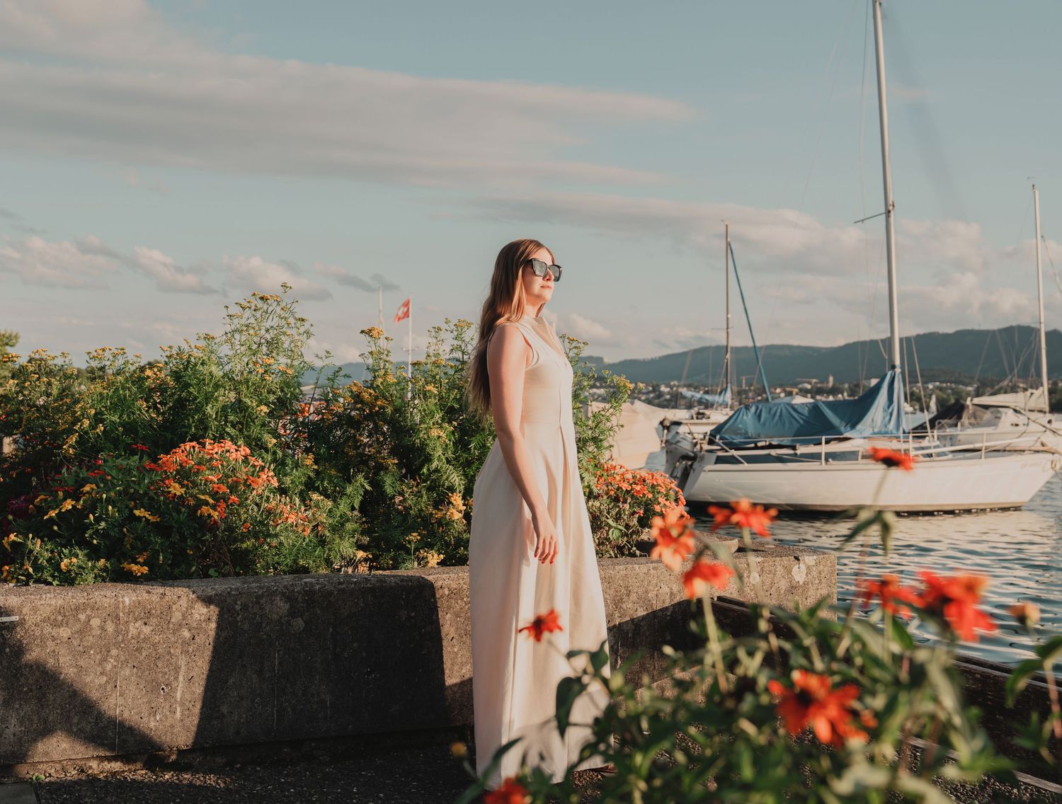 Woman in white maxi dress standing by marina with sailboats and red flowers.
