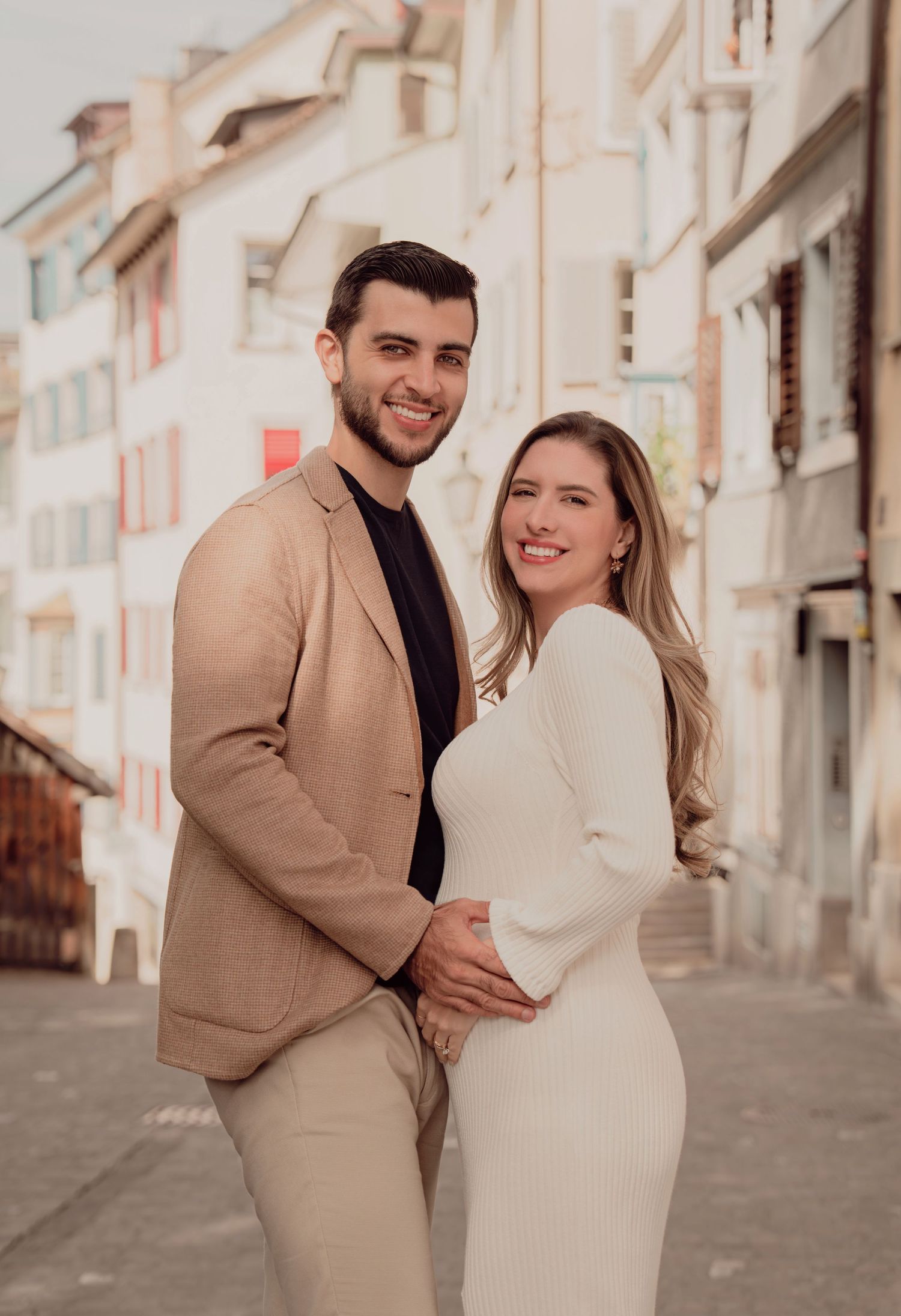 Smiling couple in coordinating beige outfits posing on a charming European cobblestone street.