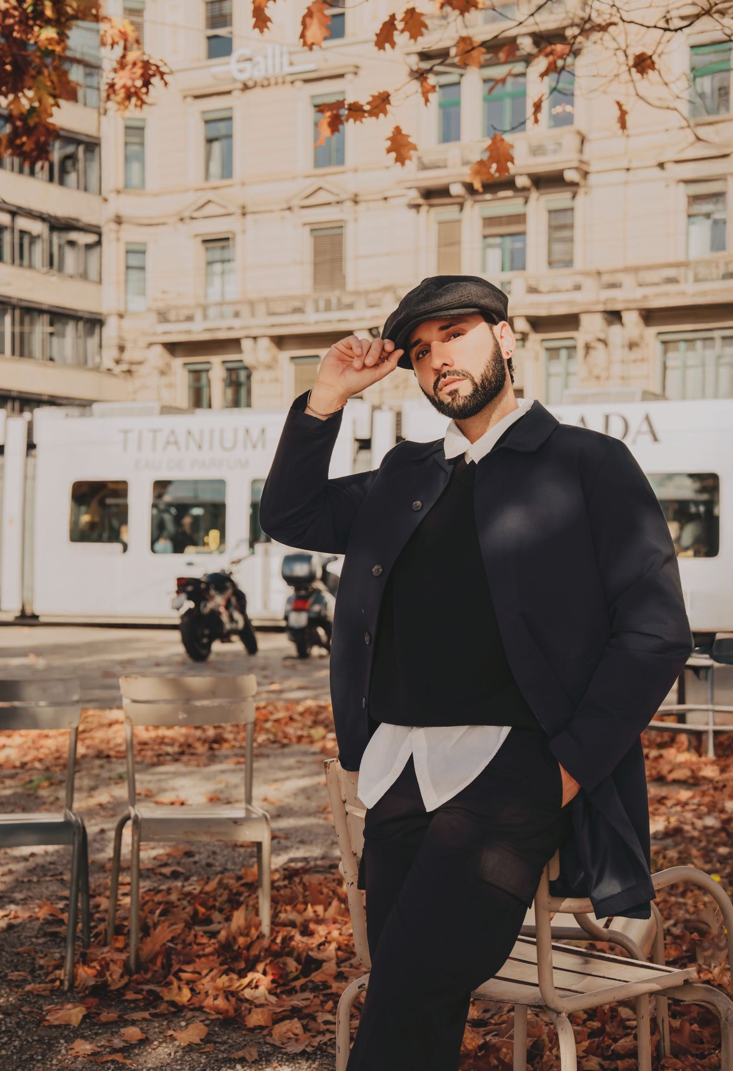 Stylish man in black coat and flat cap posing on autumn street with fallen leaves.