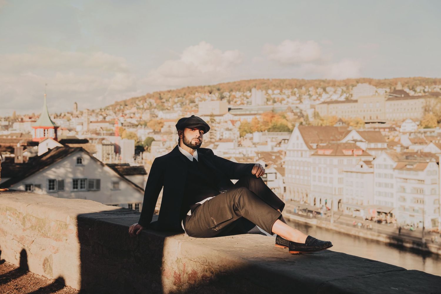 Man in black outfit sitting on a rooftop ledge overlooking a sunny European riverside cityscape.