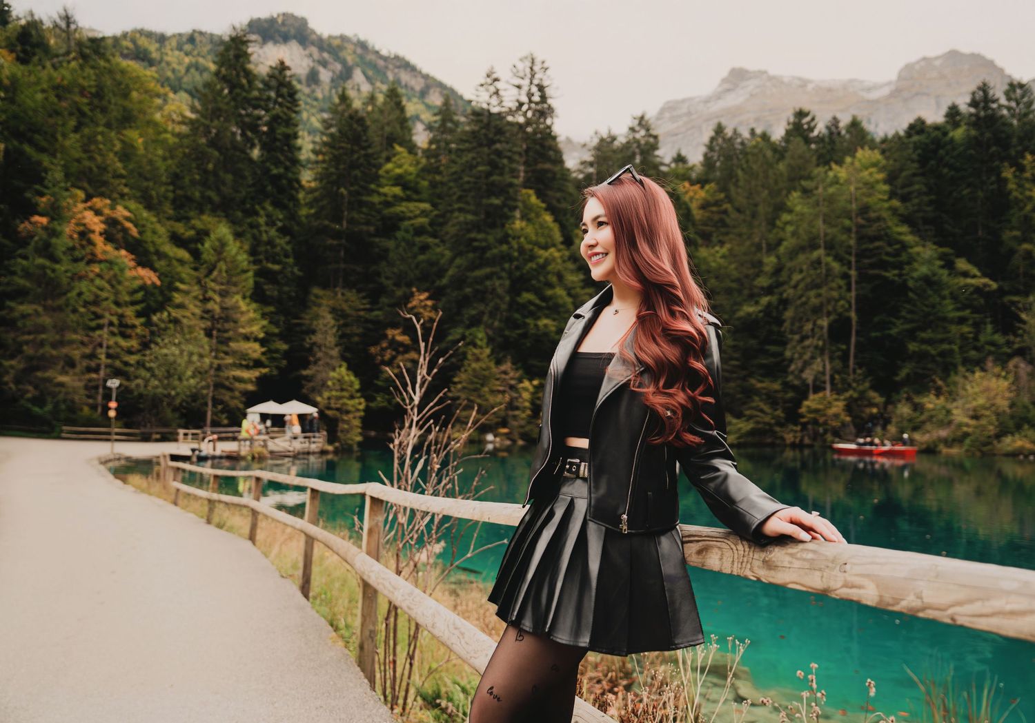 Stylish red-haired woman in black leather skirt smiling by turquoise lake with mountain views.