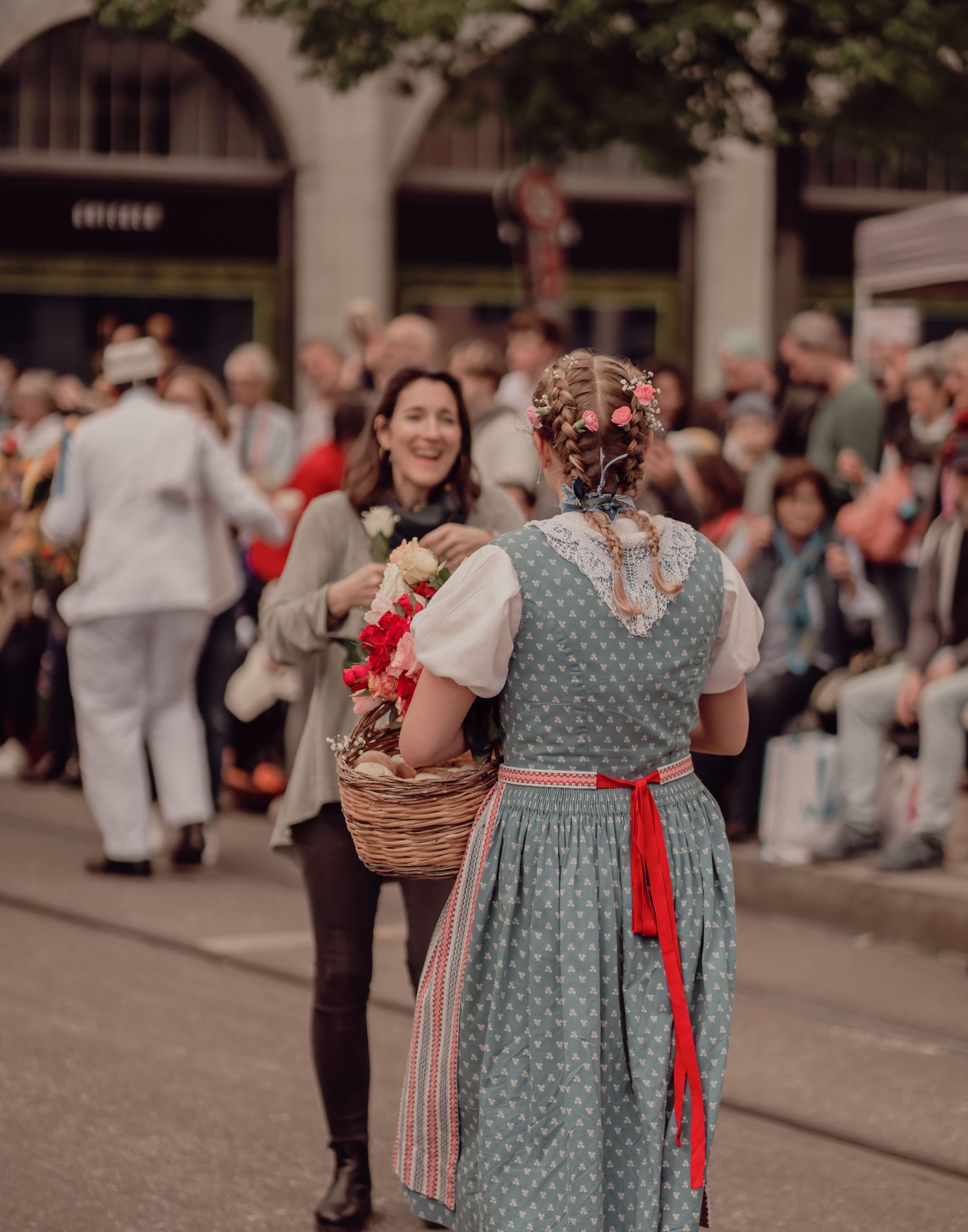 Girl in traditional dirndl dress offers flowers from basket to smiling woman at cultural parade.