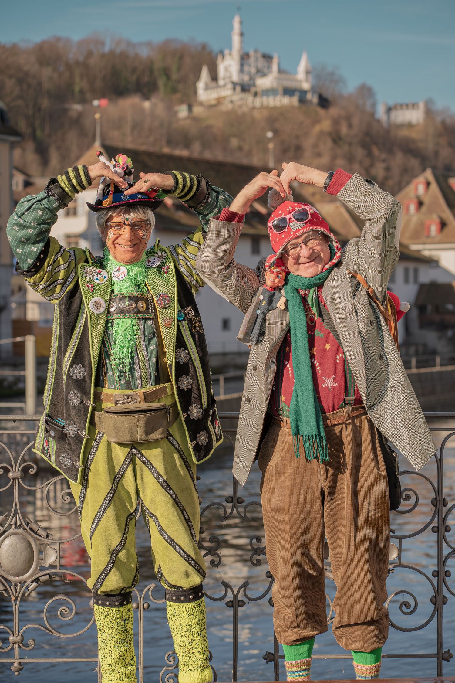 Two people in colorful carnival costumes making heart shapes with their hands on a bridge.