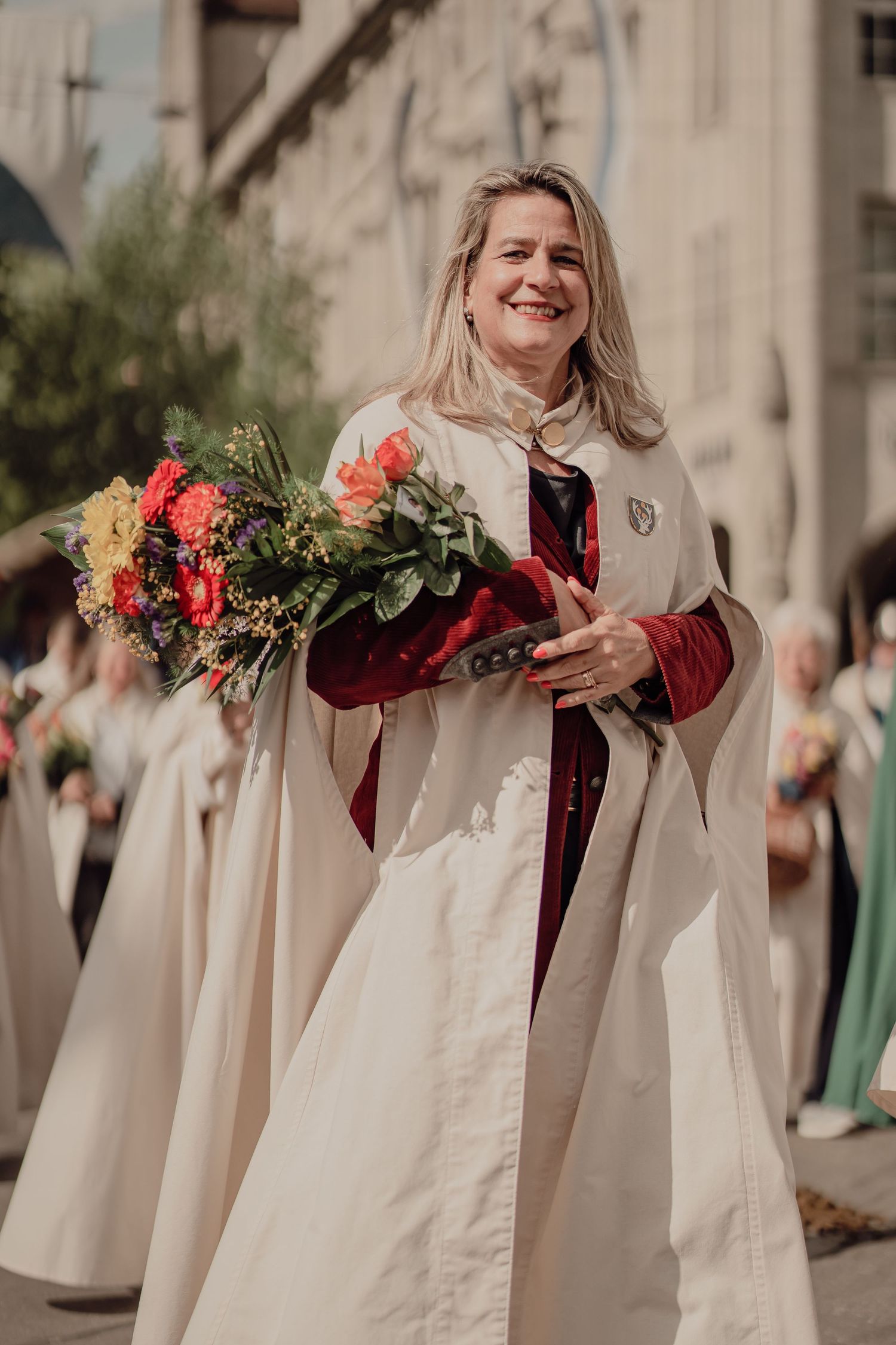Smiling woman in cream ceremonial cape holds vibrant bouquet during traditional festival procession.