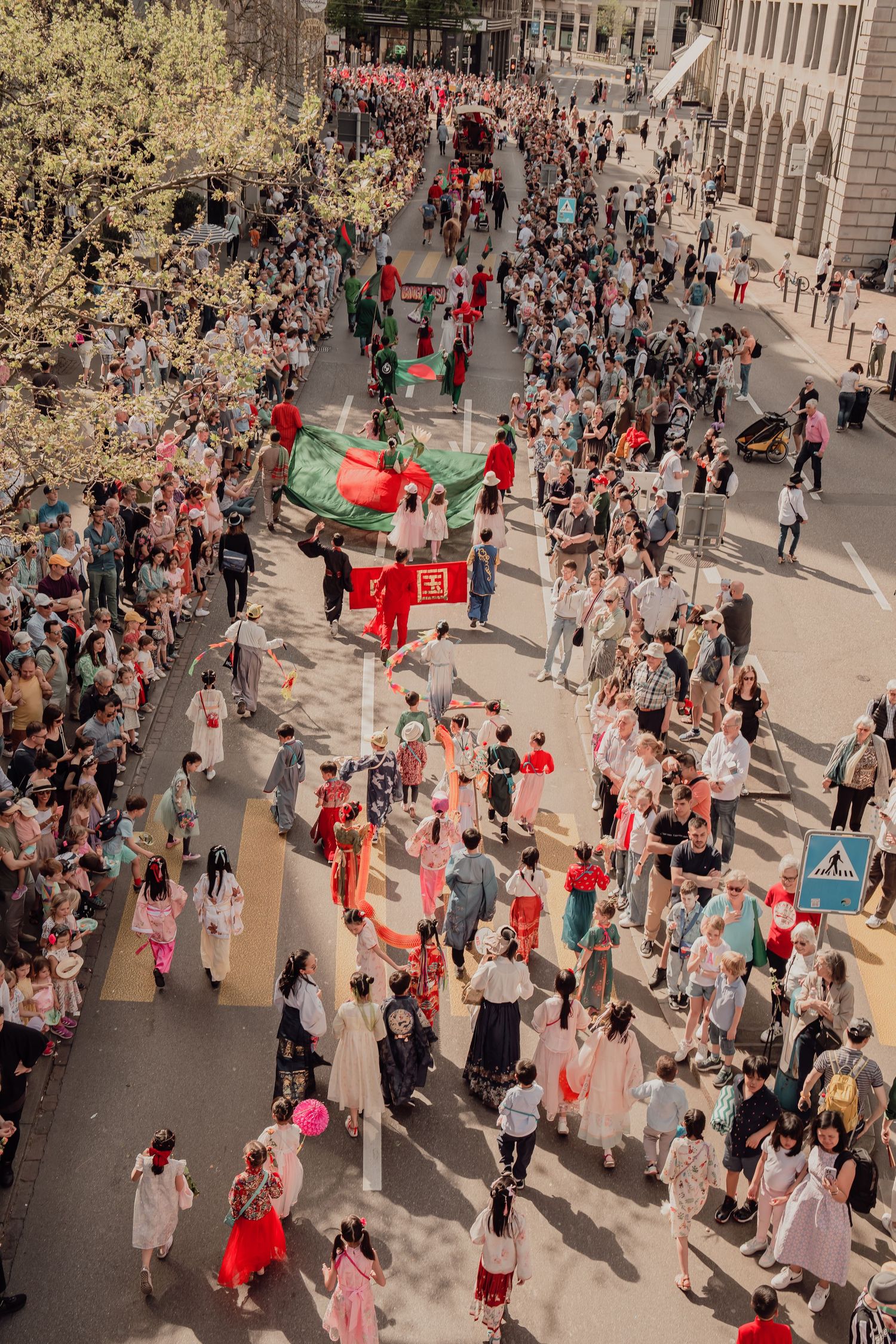 Aerial view of large festive street parade with crowds and colorful flags in city.