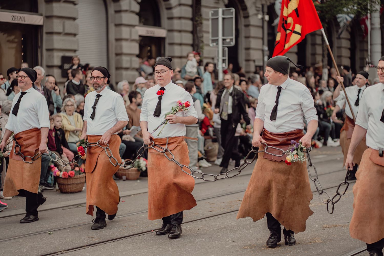 Men in leather aprons and white shirts marching in a traditional street parade.
