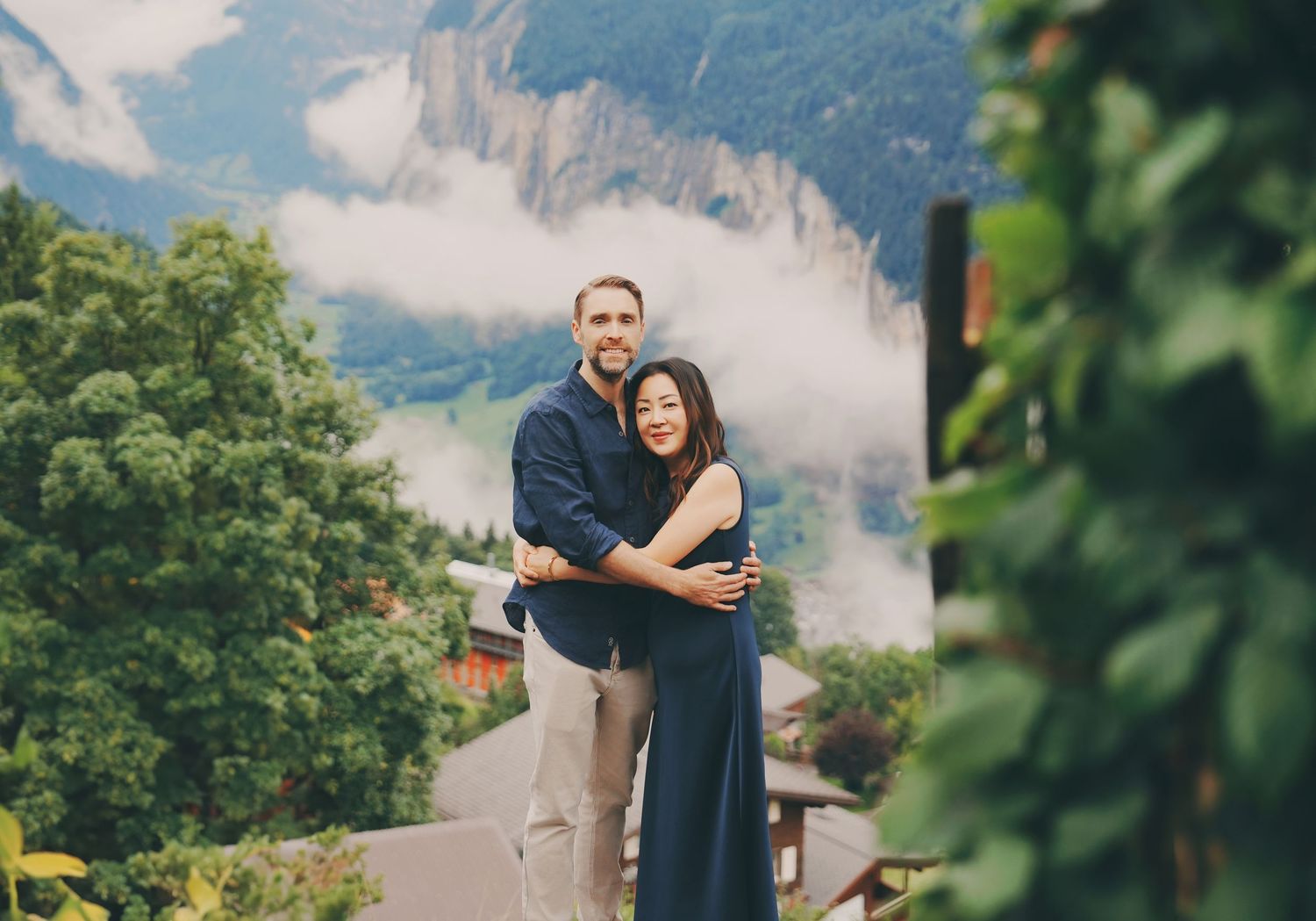 Couple smiles together on balcony surrounded by green foliage with misty Swiss mountain scenery behind.